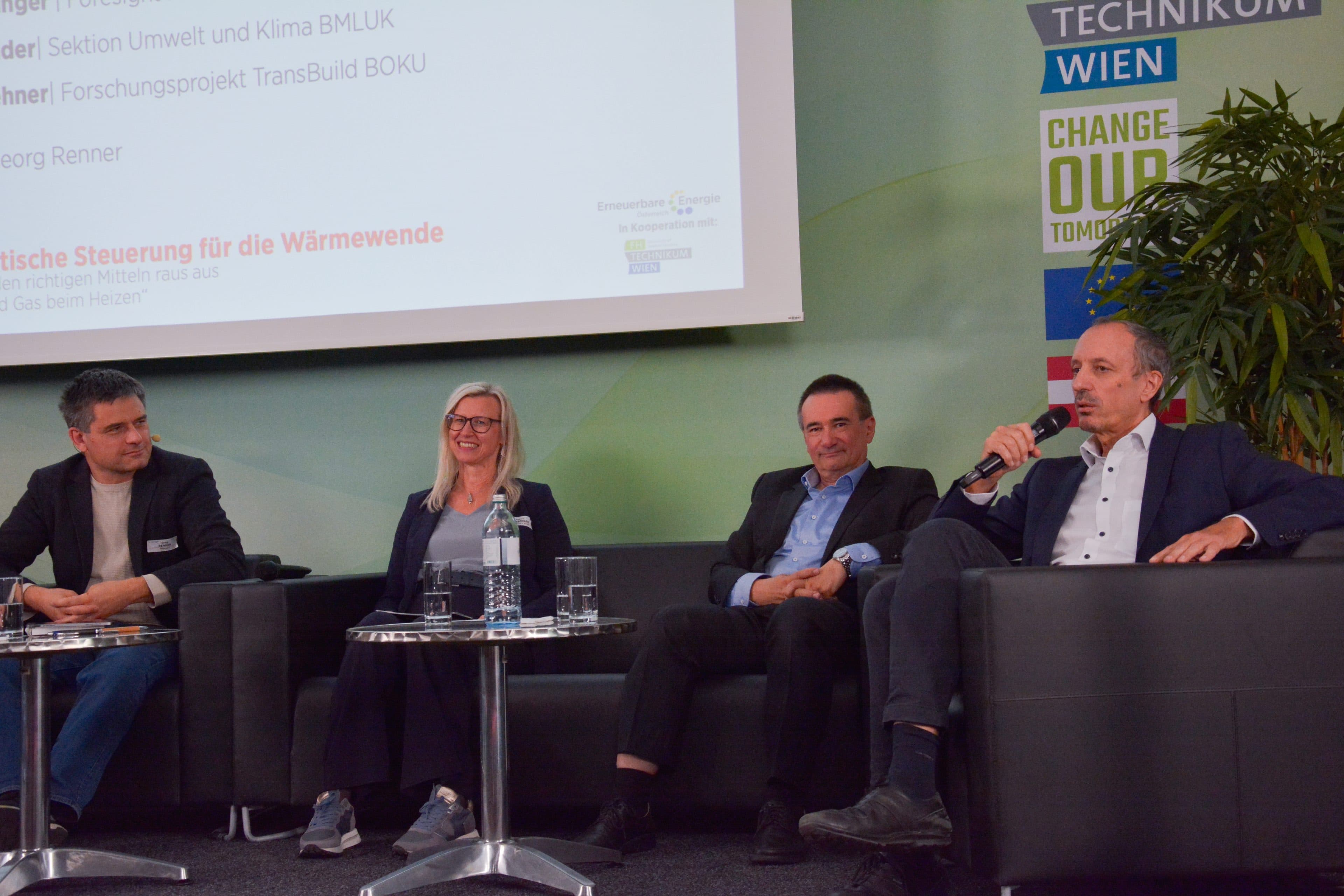 Vier Personen sitzen während einer Podiumsdiskussion in Sesseln auf einer Bühne; ein Redner hält ein Mikrofon in der Hand, im Hintergrund ist eine Präsentationsleinwand zu sehen.