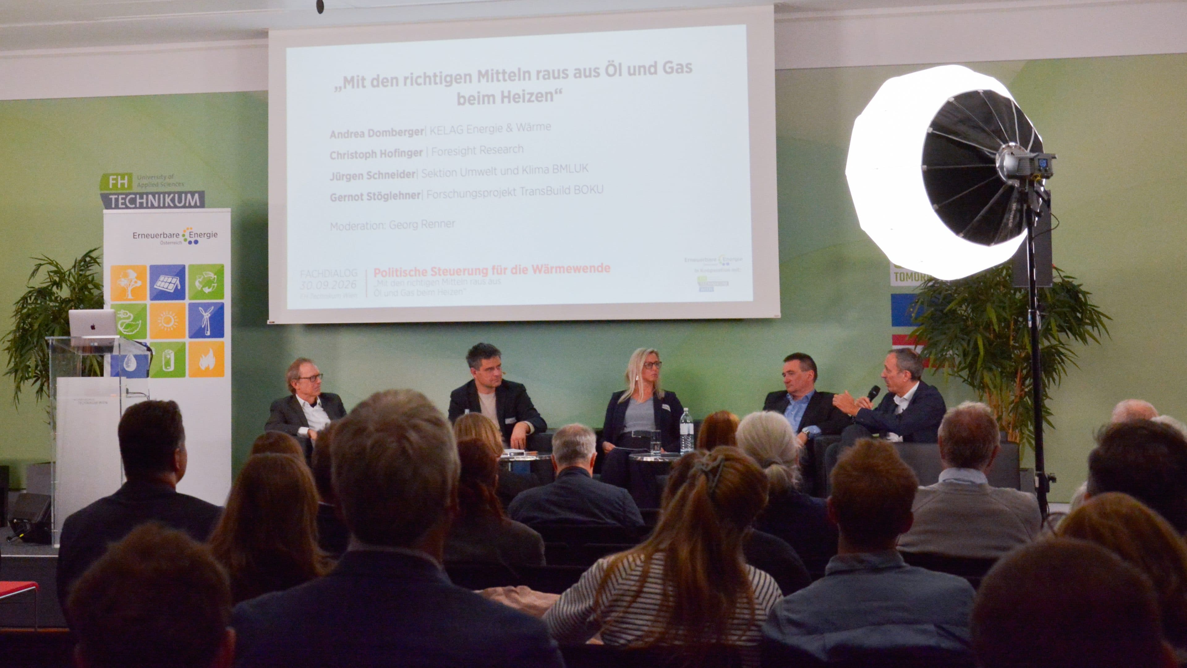 Eine Diskussionsrunde auf der Bühne bei einer Veranstaltung, hinter der auf einer Leinwand Informationen zum Umstieg von Öl- und Gasheizungen angezeigt werden.