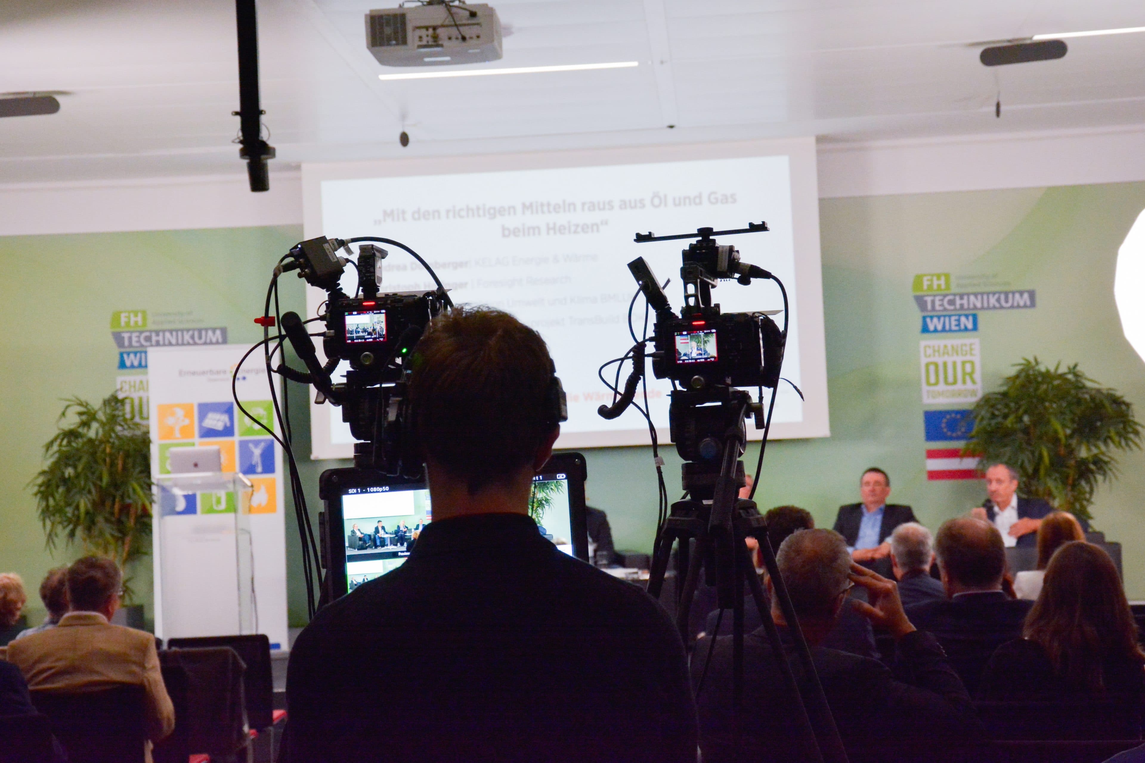 Eine Person bedient eine Videokamera, die auf eine Präsentationsleinwand und eine Podiumsrunde bei einer Konferenz gerichtet ist.