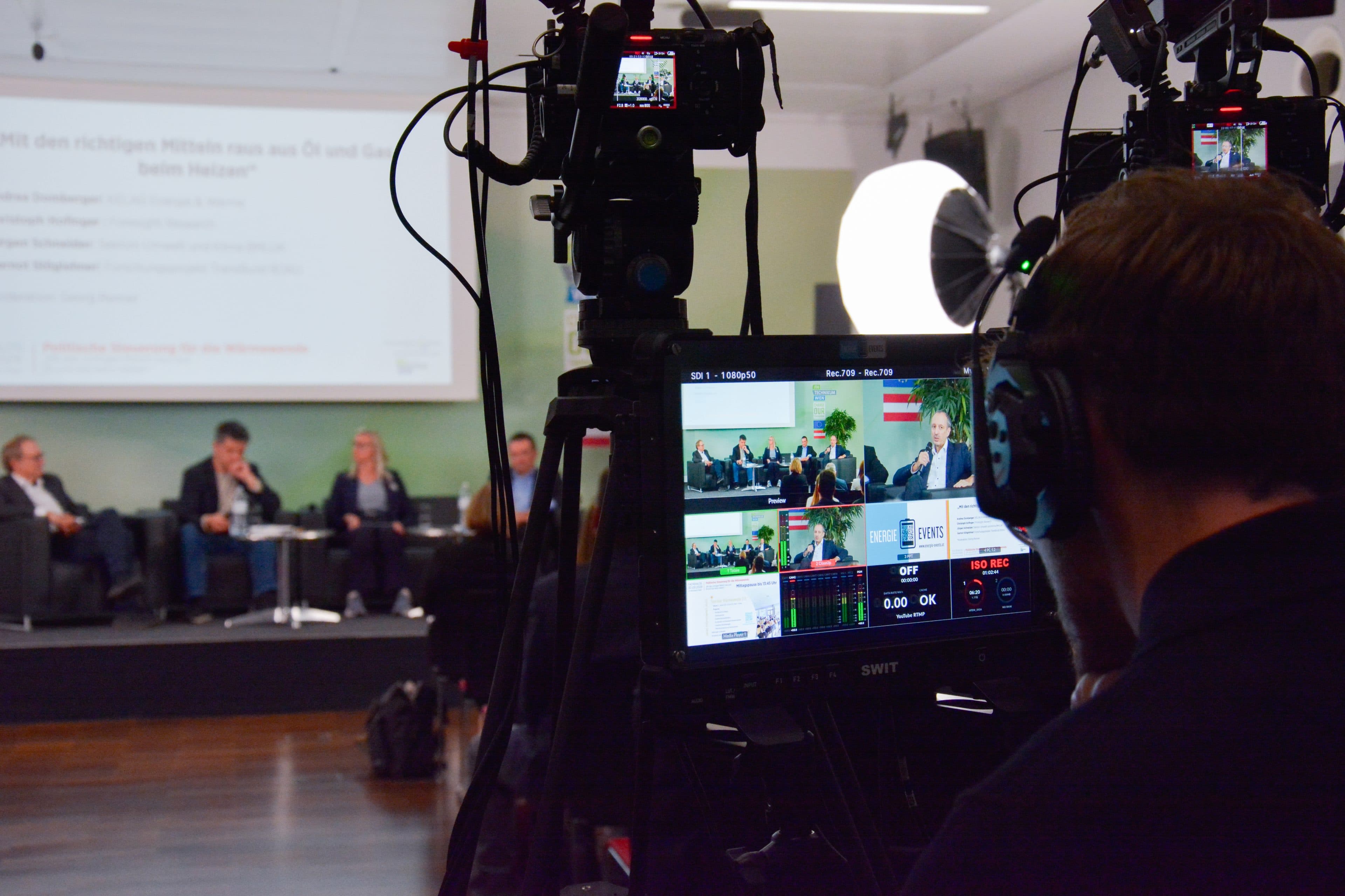 Ein Kameramann filmt eine Podiumsdiskussion auf einer deutschsprachigen Konferenz. Auf einem Monitor ist das Live-Bild zu sehen. Die Podiumsteilnehmer sitzen auf der Bühne vor einer projizierten Präsentationsfolie.