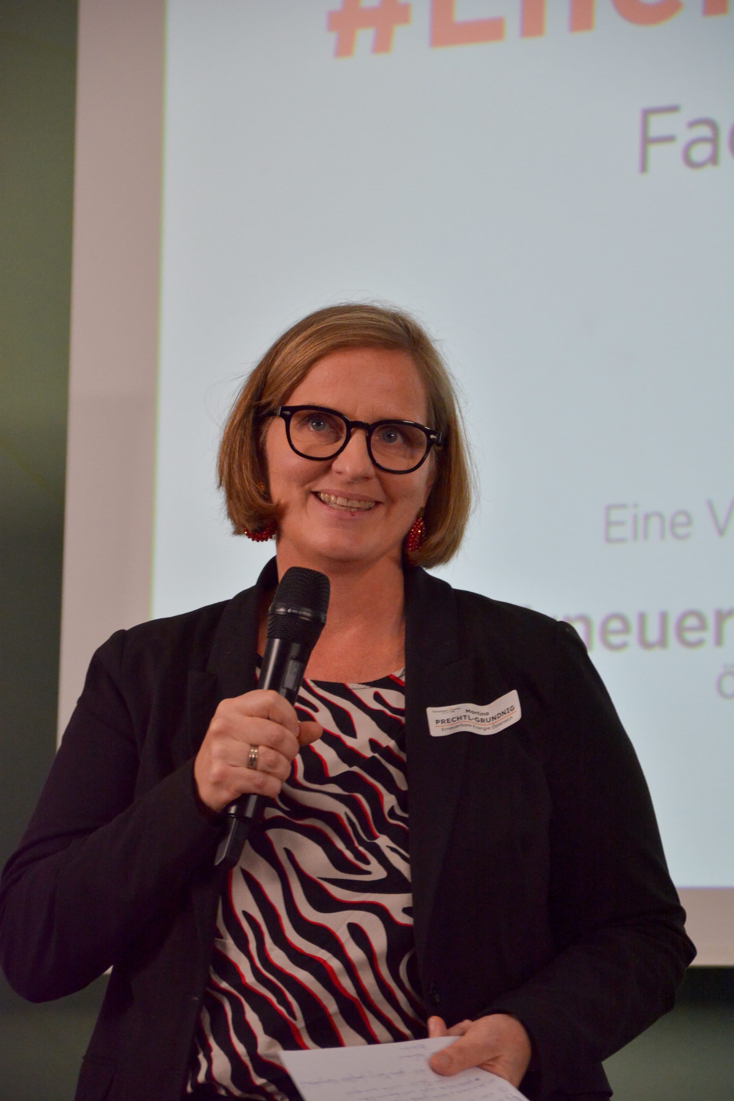 Eine Frau mit Brille und Blazer spricht vor einer Leinwand in ein Mikrofon.