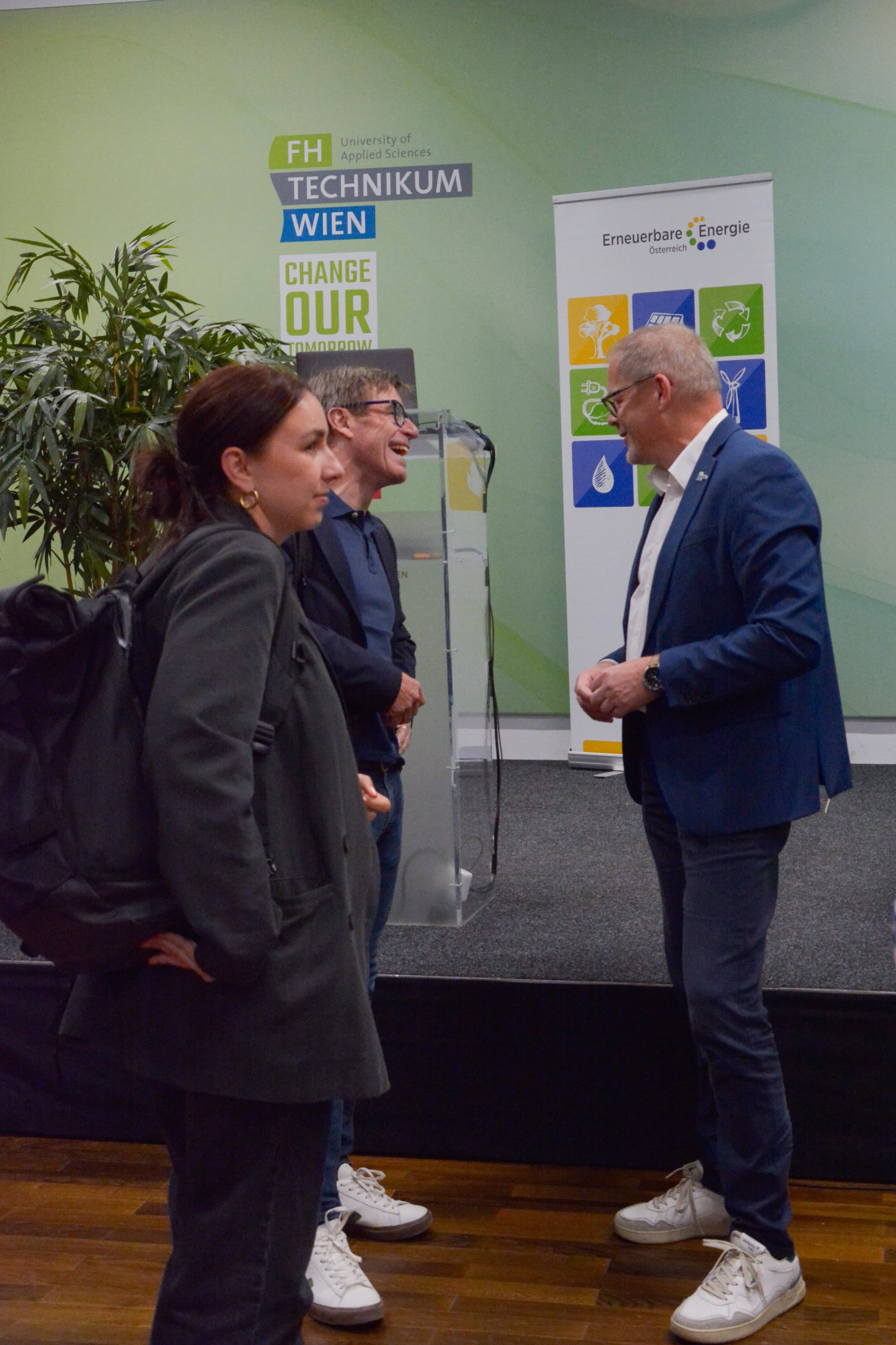 Drei Personen stehen auf einer Konferenz vor einer Kulisse mit den Logos des FH Technikum Wien und von Erneuerbare Energie Österreich und unterhalten sich.