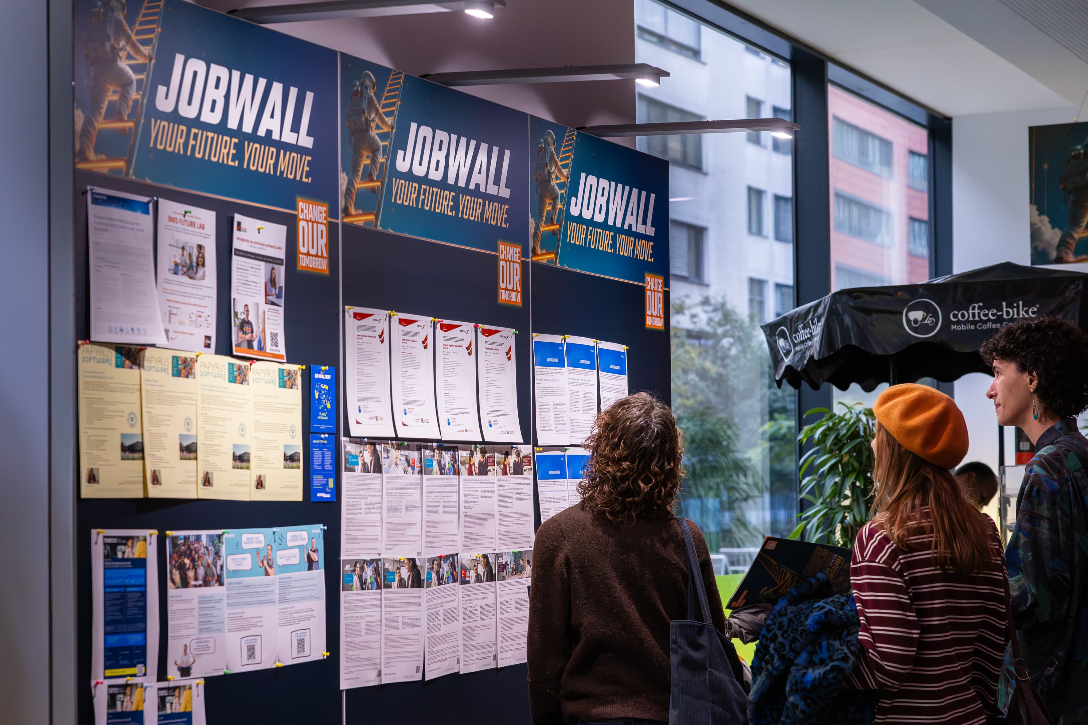Zwei Personen stehen vor einer großen Jobbörse mit der Aufschrift Jobwall: Deine Zukunft. Dein Schritt, die mit verschiedenen ausgedruckten Stellenanzeigen bedeckt ist.