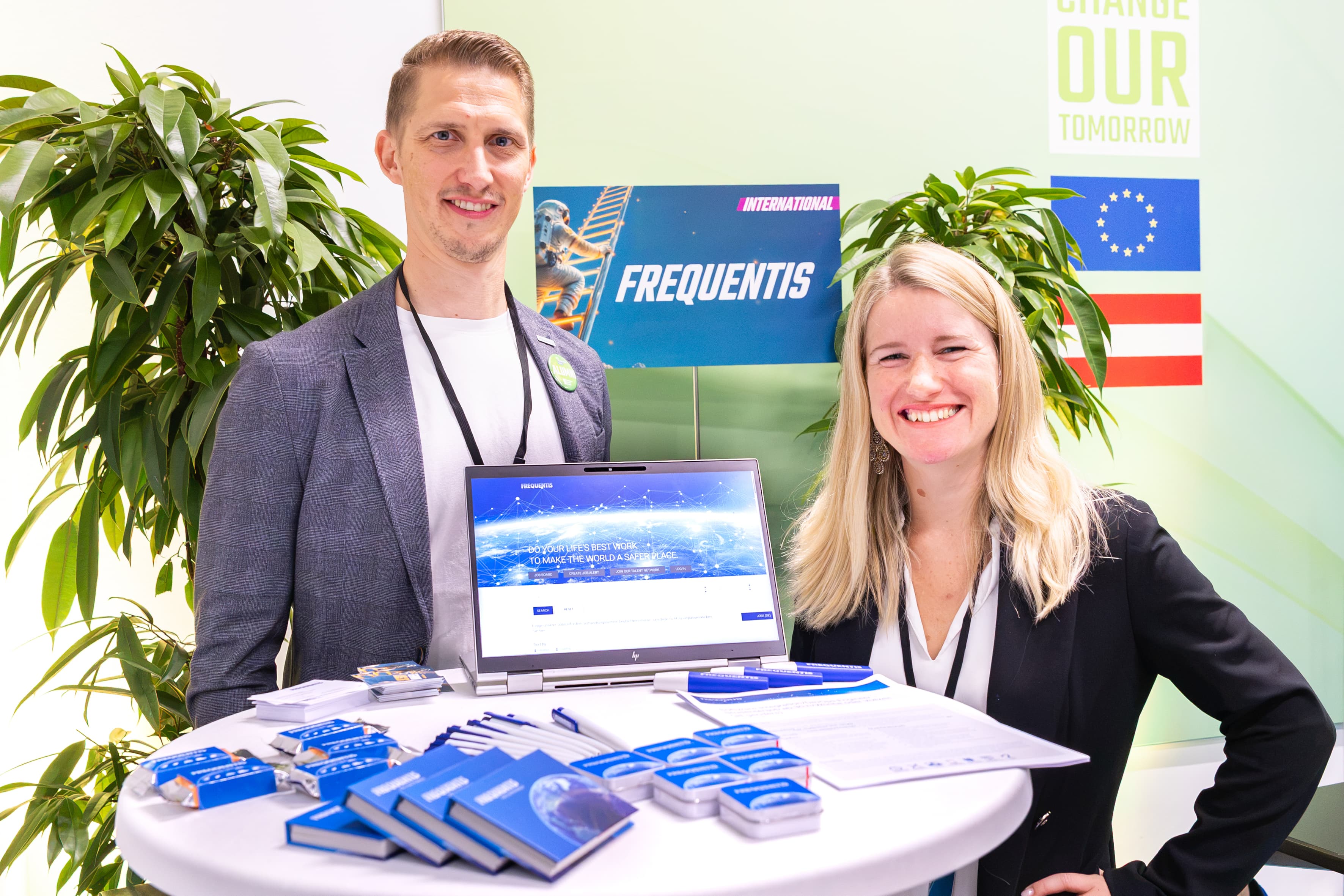 Ein Mann und eine Frau stehen hinter einem Messestand, auf dem ein Laptop steht, auf dem die Website von Frequentis zu sehen ist, sowie Werbematerialien mit dem Firmenlogo.