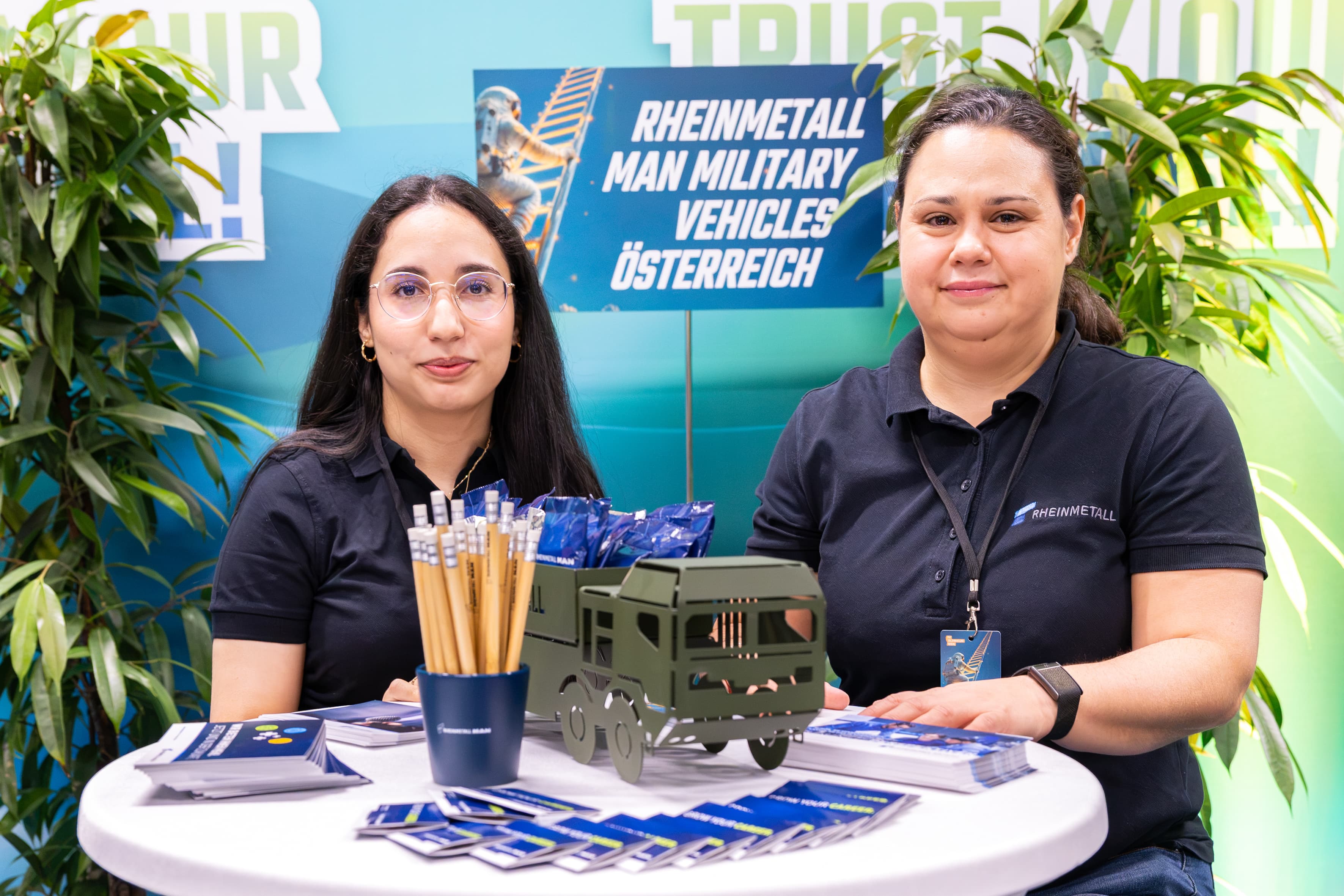 Zwei Frauen stehen hinter einem Stand von Rheinmetall MAN Military Vehicles, auf dem Werbematerialien, ein Modell eines Militär-Lkw und eine Schale mit Bleistiften ausgestellt sind.