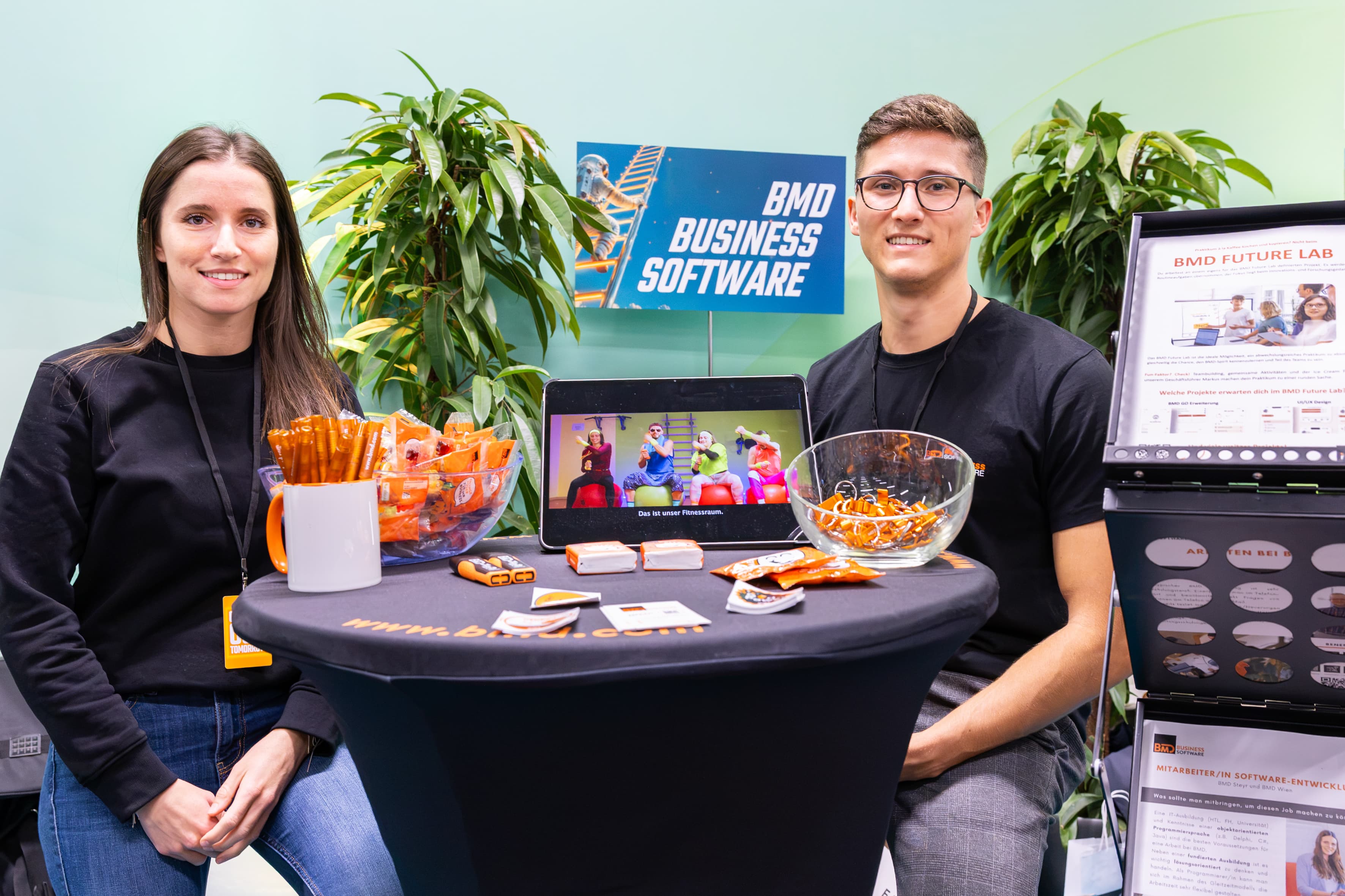 Ein Mann und eine Frau stehen hinter einem Tisch am Stand von BMD Business Software; auf einem Tablet wird ein Fitnessvideo gezeigt, und auf dem Tisch liegen verschiedene Werbeartikel.