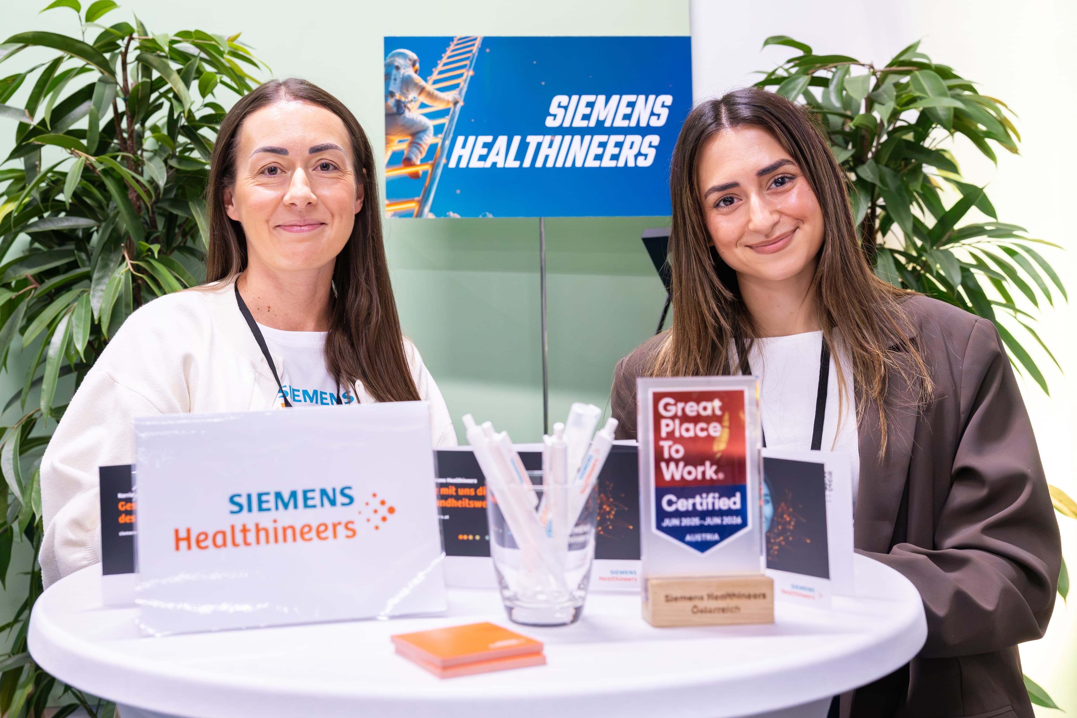 Zwei Frauen stehen an einem Informationsstand der Marke Siemens Healthineers, auf dessen Tisch eine Great Place to Work-Auszeichnung steht.