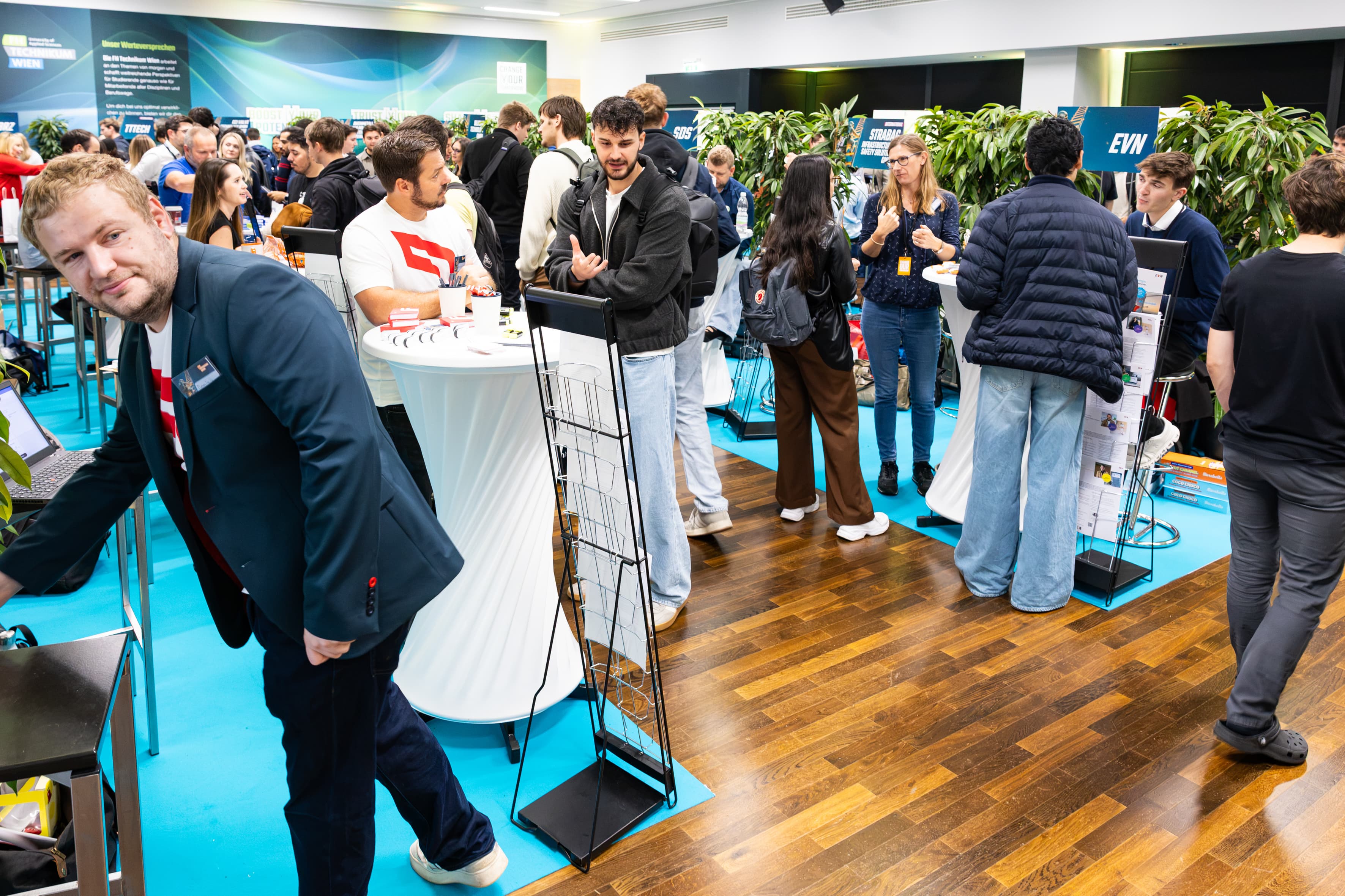 Menschen, die auf einer Jobmesse mit Unternehmensständen Kontakte knüpfen und an Stehtischen auf einem blauen Boden stehen.