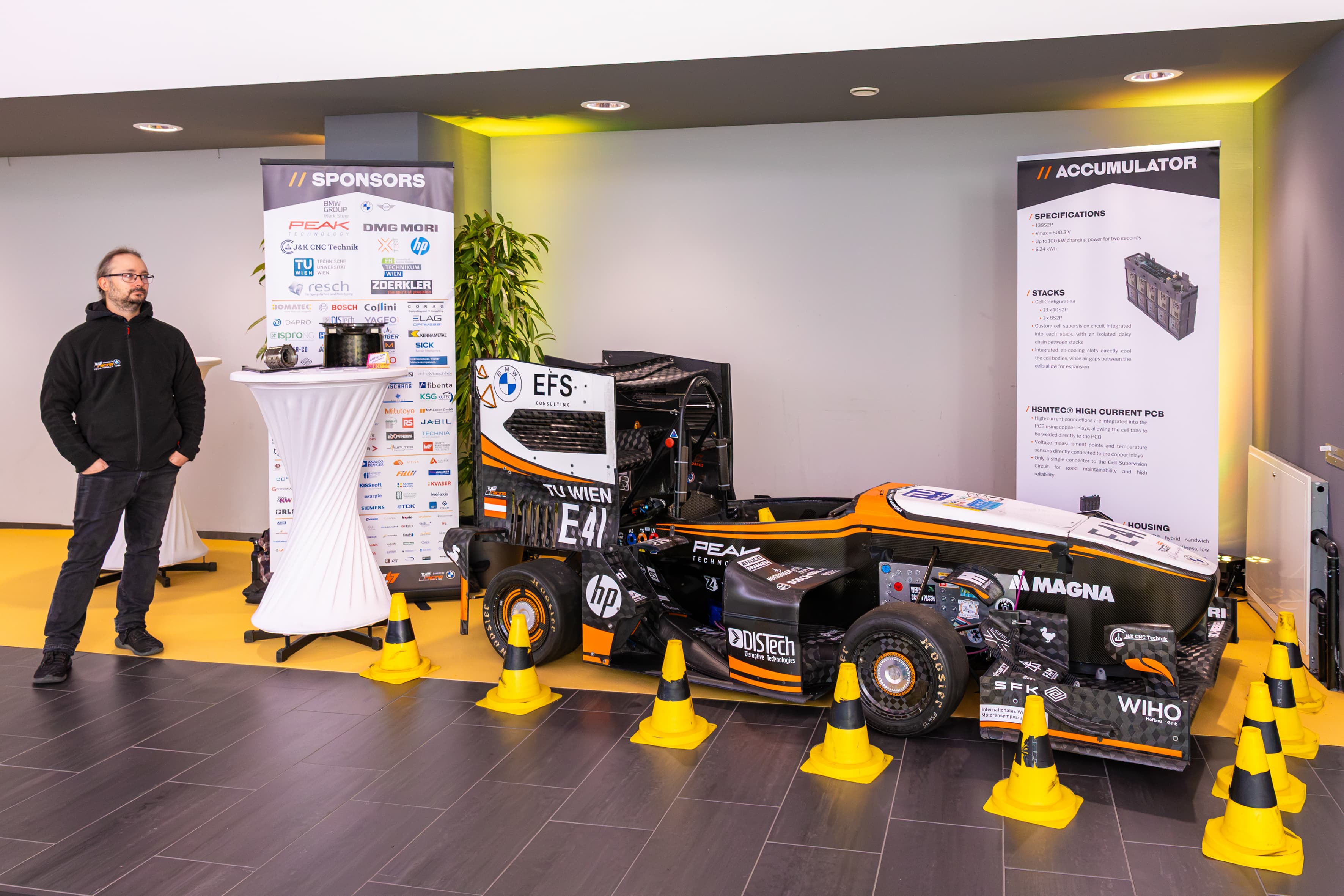 Ein Mann steht neben einer Ausstellung, die einen Formula Student-Rennwagen der TU Wien, ein Sponsorenbanner und eine Informationstafel zum Thema Akkumulatoren zeigt.