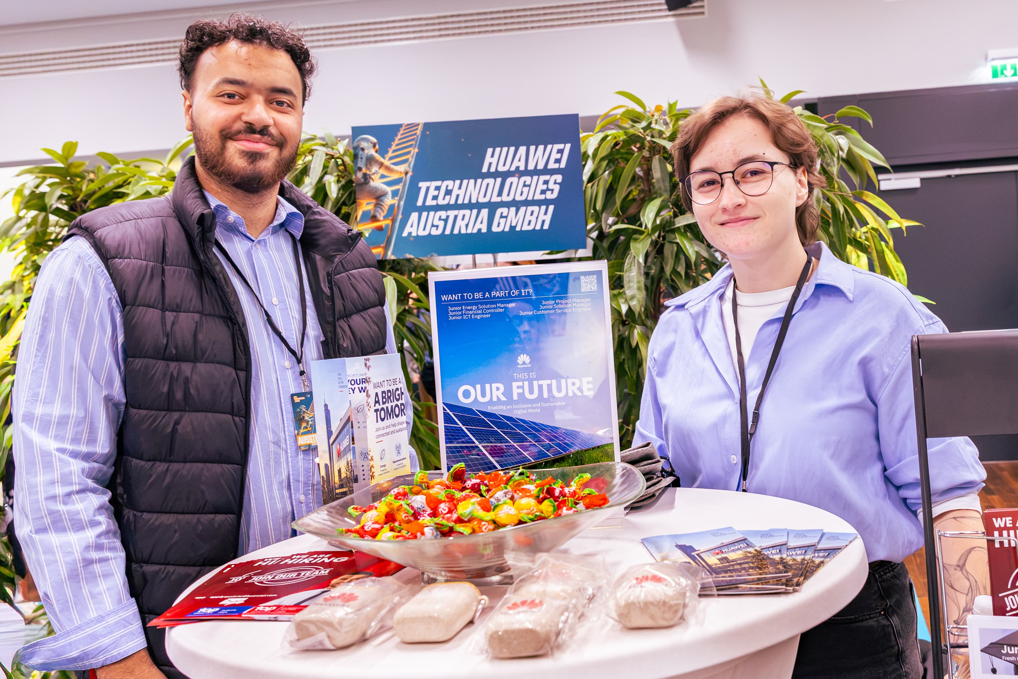 Hinter einem Tisch am Stand der Huawei Technologies Austria GmbH stehen zwei Personen mit einer Schale voller verpackter Bonbons und Broschüren zur Personalrekrutierung.
