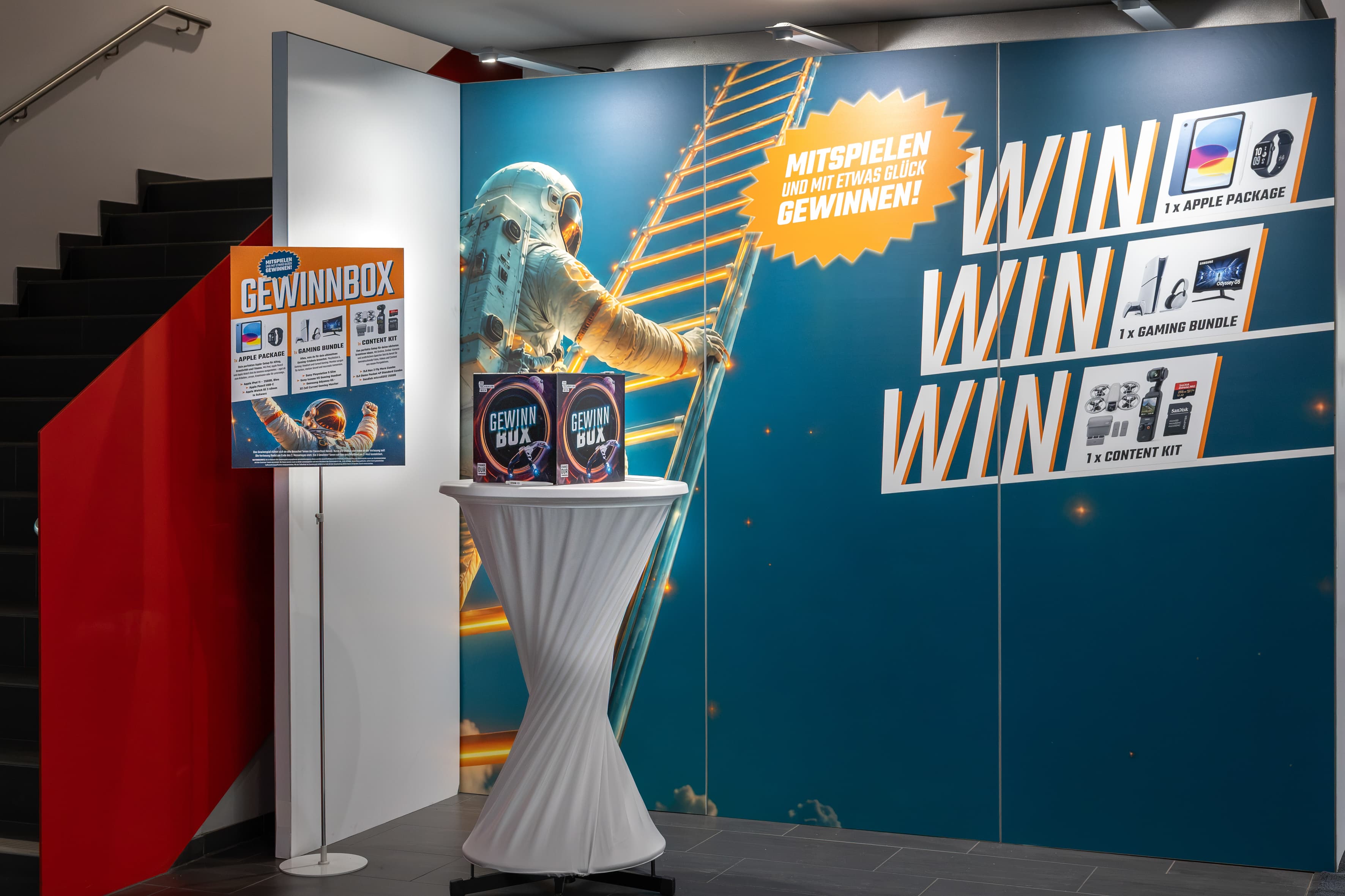 Ein Werbedisplay, auf dem ein Astronaut eine Leiter hinaufklettert, mit Informationen zu den Preisen: ein Apple-Paket, ein Gaming-Bundle und ein Content-Kit, einschließlich eines Schildes und von Kisten mit der Aufschrift Gewinnbox.