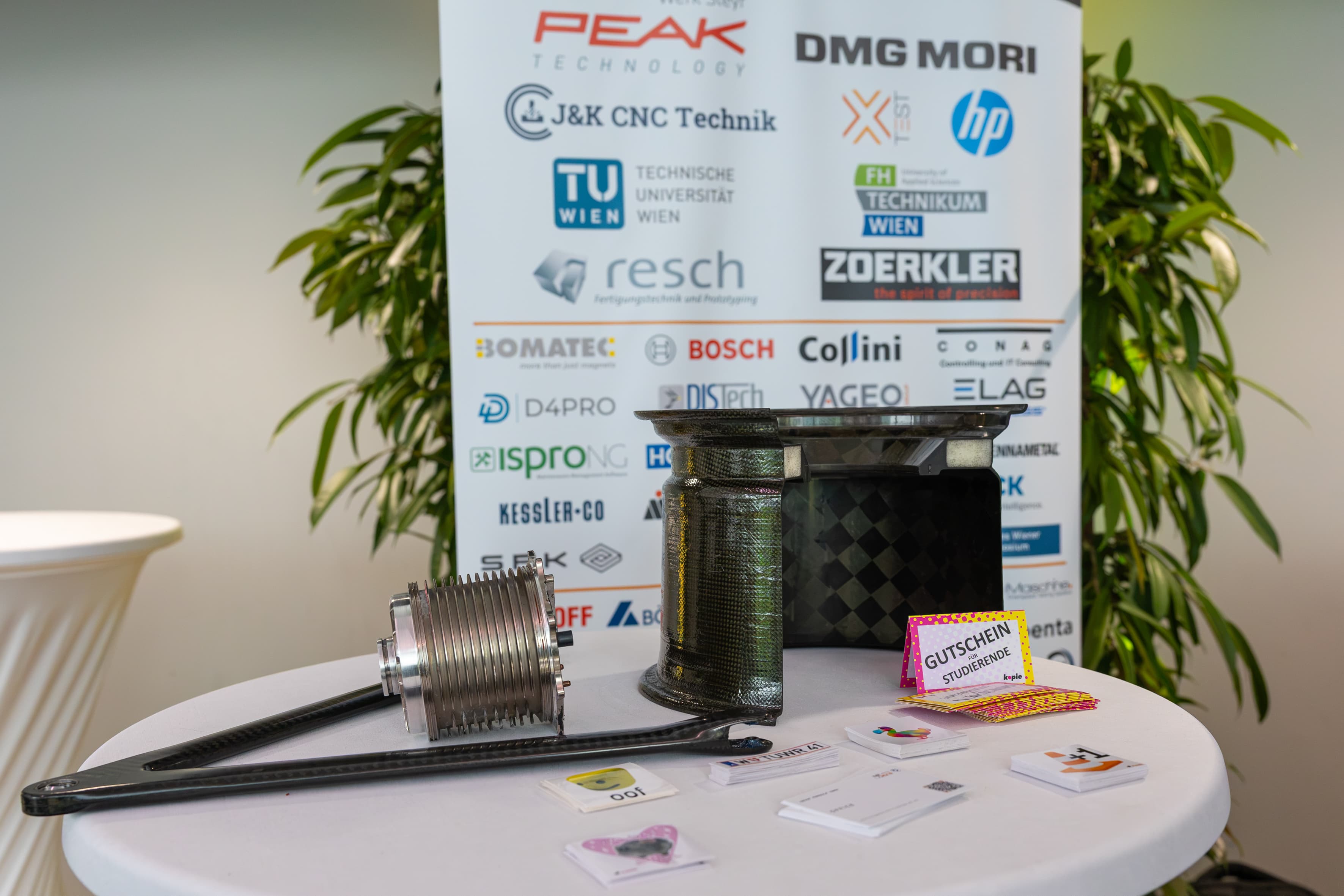Ein mechanisches Bauteil und eine Felge aus Kohlefaser, ausgestellt auf einem weißen Tisch vor einem Banner eines Unternehmenssponsors.