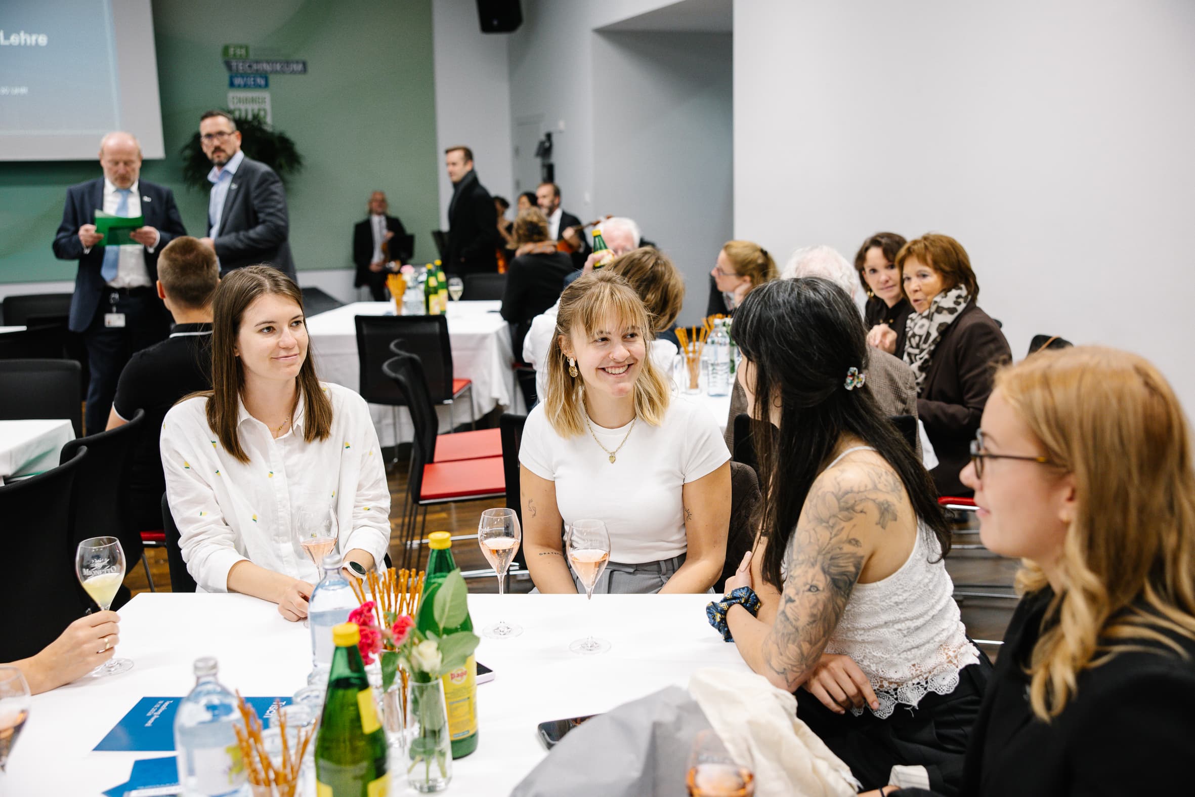 Bei einer Veranstaltung sitzt eine Gruppe von Menschen um einen Tisch herum; im Vordergrund sind mehrere Frauen zu sehen, die lächelnd miteinander reden und dabei Weingläser in der Hand halten.