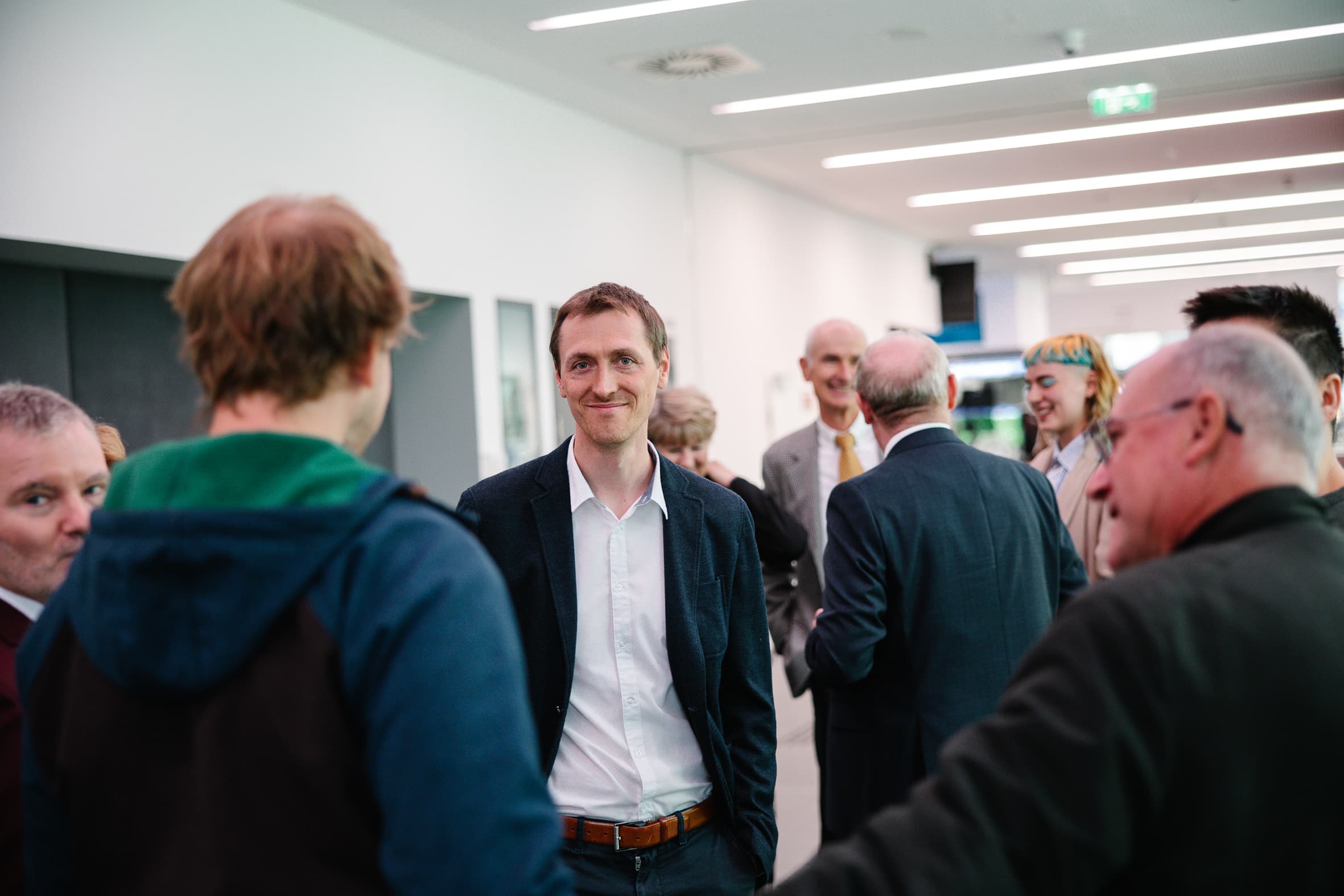 Ein Mann in Blazer und weißem Hemd steht in einem Flur und unterhält sich lächelnd mit einer Gruppe von Menschen.