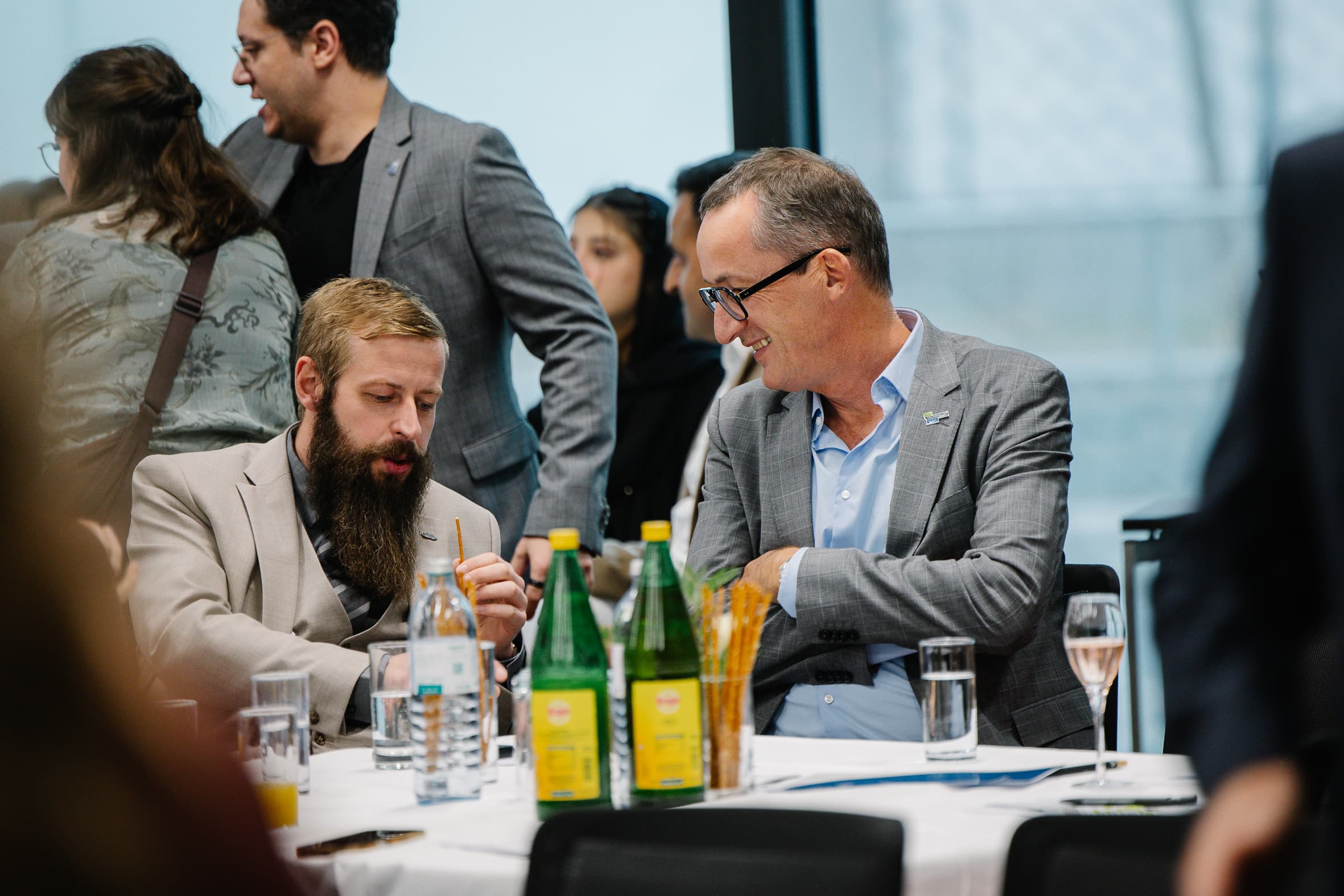 Zwei Männer in Business-Casual-Kleidung sitzen an einem Konferenztisch, unterhalten sich und lächeln, während andere Teilnehmer im Hintergrund stehen.