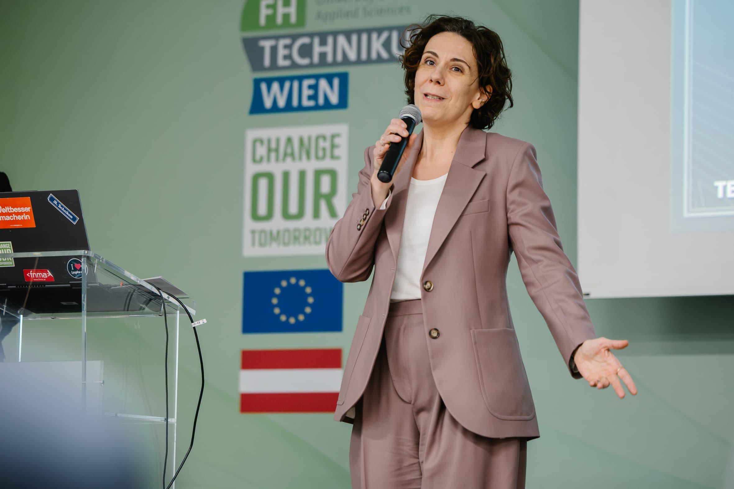 Eine Frau in einem mauvefarbenen Sakko spricht in ein Mikrofon und gestikuliert dabei in Richtung des Publikums.