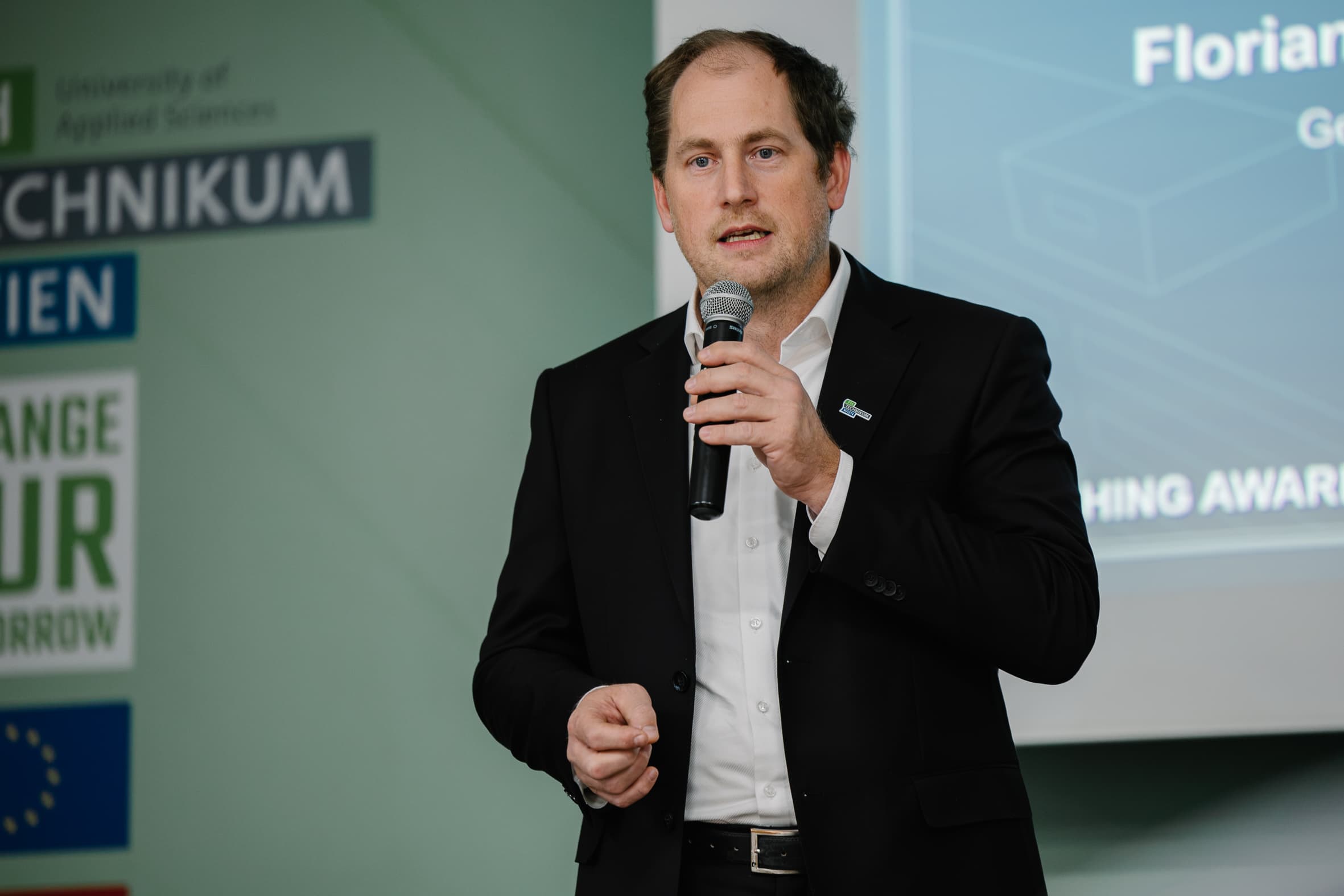 Ein Mann in einem schwarzen Sakko und einem weißen Hemd spricht vor einer Präsentationsleinwand in ein Mikrofon.