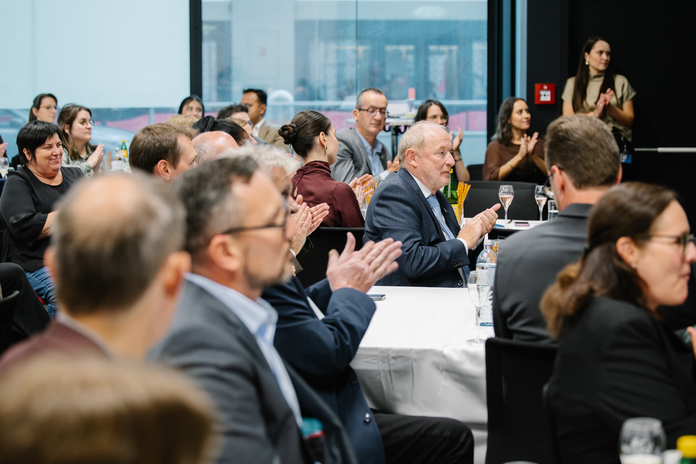 Menschen, die an runden Tischen in einem Konferenzraum sitzen und während einer Präsentation klatschen.