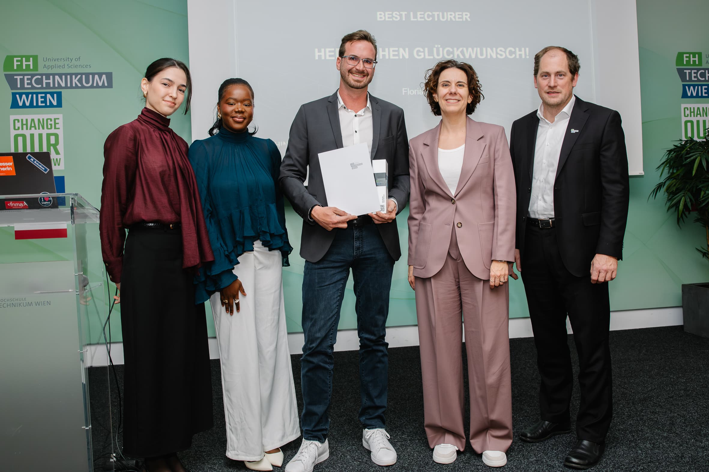 Fünf Personen stehen bei einer Preisverleihung in einer Reihe, wobei die Person in der Mitte eine Urkunde vor einem Banner mit der Aufschrift Bester Dozent in der Hand hält.