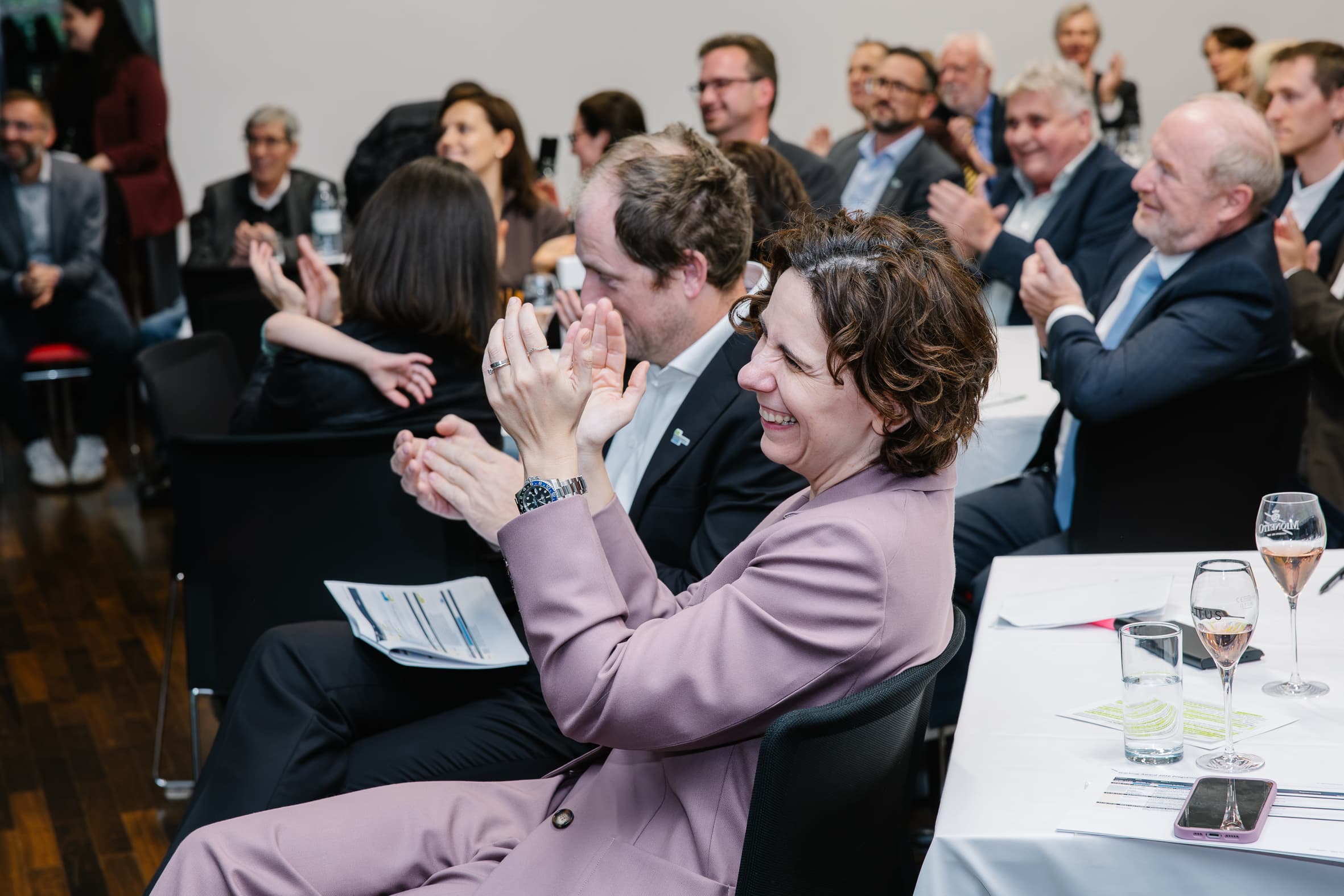 Eine Frau in einem hellvioletten Kostüm sitzt bei einer Konferenz mit anderen Teilnehmern an einem Tisch, lächelt und klatscht.