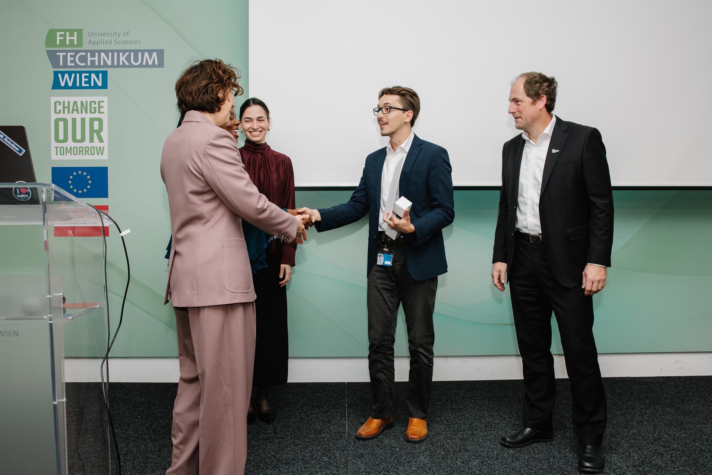 Eine Frau in einem lila Kostüm schüttelt einem Mann die Hand, der eine kleine weiße Schachtel in der Hand hält, während eine weitere Frau und ein Mann in einem schwarzen Anzug vor einem Banner der Fachhochschule Technikum Wien zuschauen.