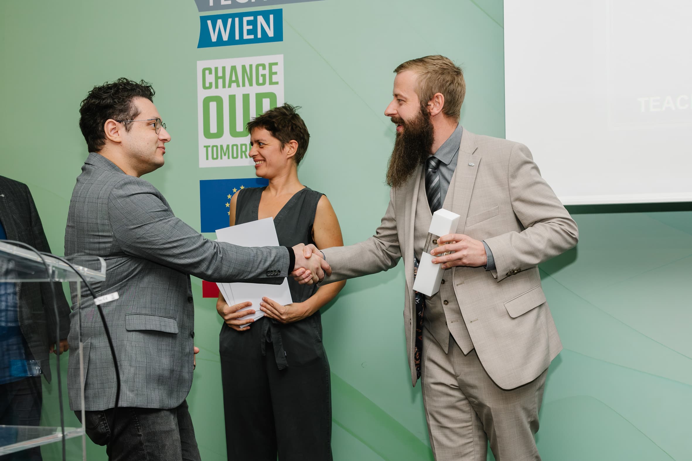 Ein Mann in einem grauen Anzug gibt einem Mann in einem grauen Blazer die Hand, während eine Frau in einem dunklen Overall zwischen ihnen steht und Papiere in der Hand hält.
