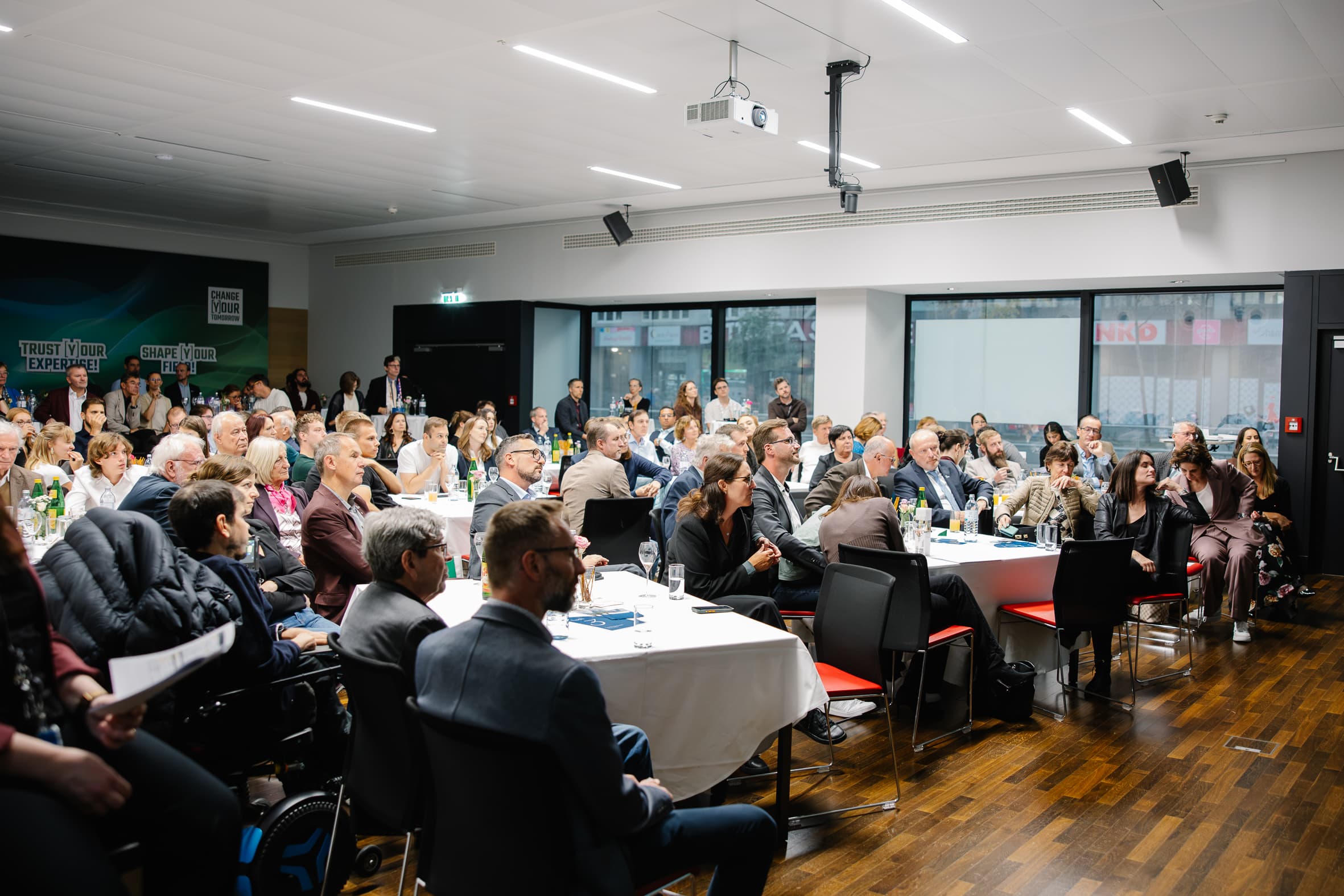 Während einer Präsentation sitzt das Publikum an runden Tischen in einem Konferenzraum.