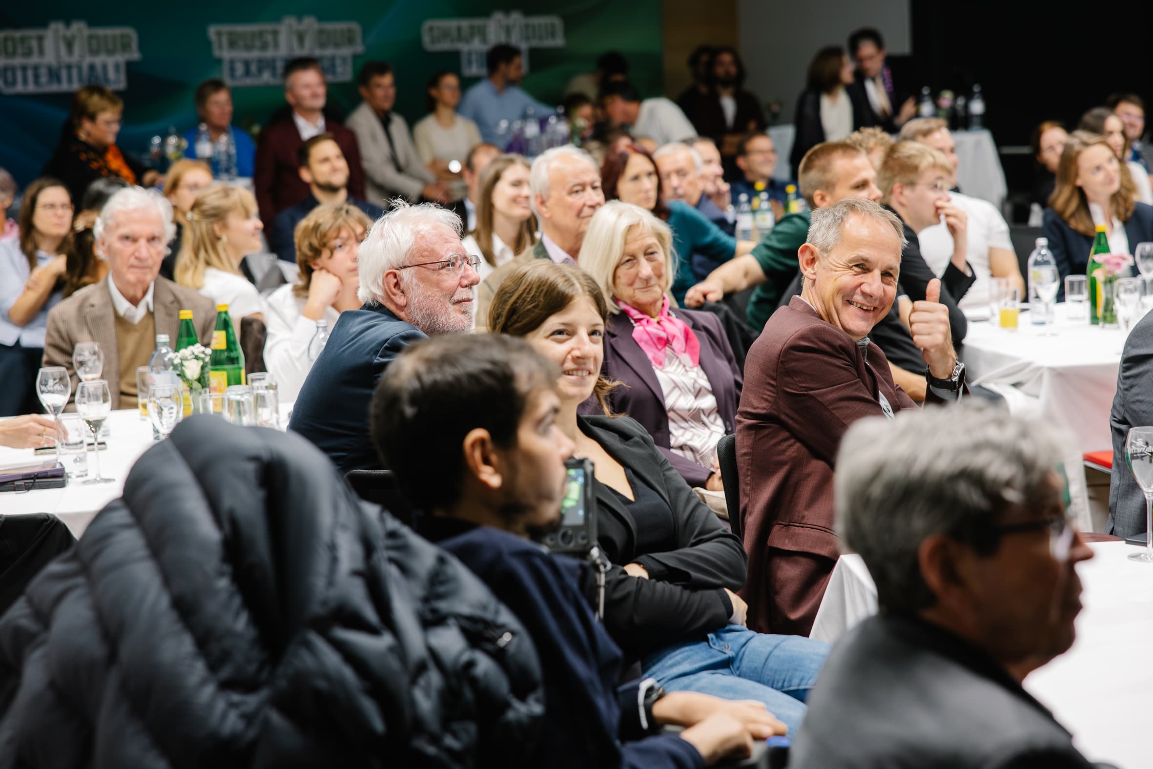 Eine Gruppe von Menschen, die während einer Konferenz an Tischen sitzen; im Vordergrund ist ein Mann zu sehen, der den Daumen hochhält.