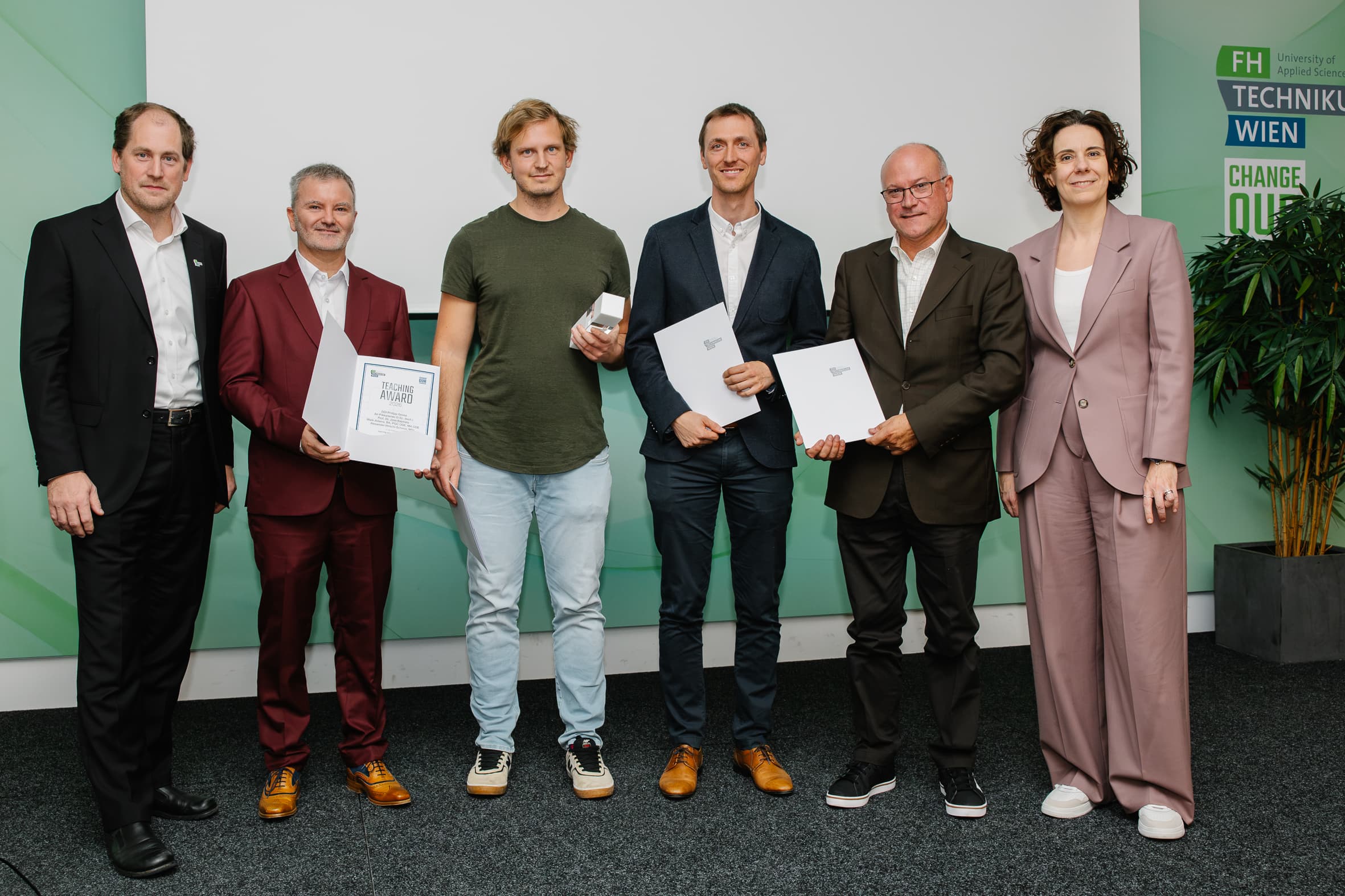 Sechs Personen stehen bei einer Preisverleihung in einer Reihe, wobei drei von ihnen Urkunden und eine Person eine kleine Trophäenschachtel in der Hand hält.