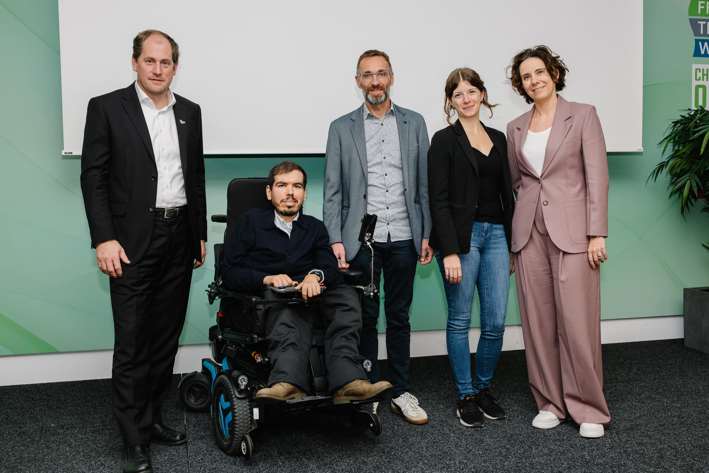 Fünf Personen stehen in einer Reihe auf einer Bühne, darunter eine Person, die in einem elektrischen Rollstuhl sitzt.