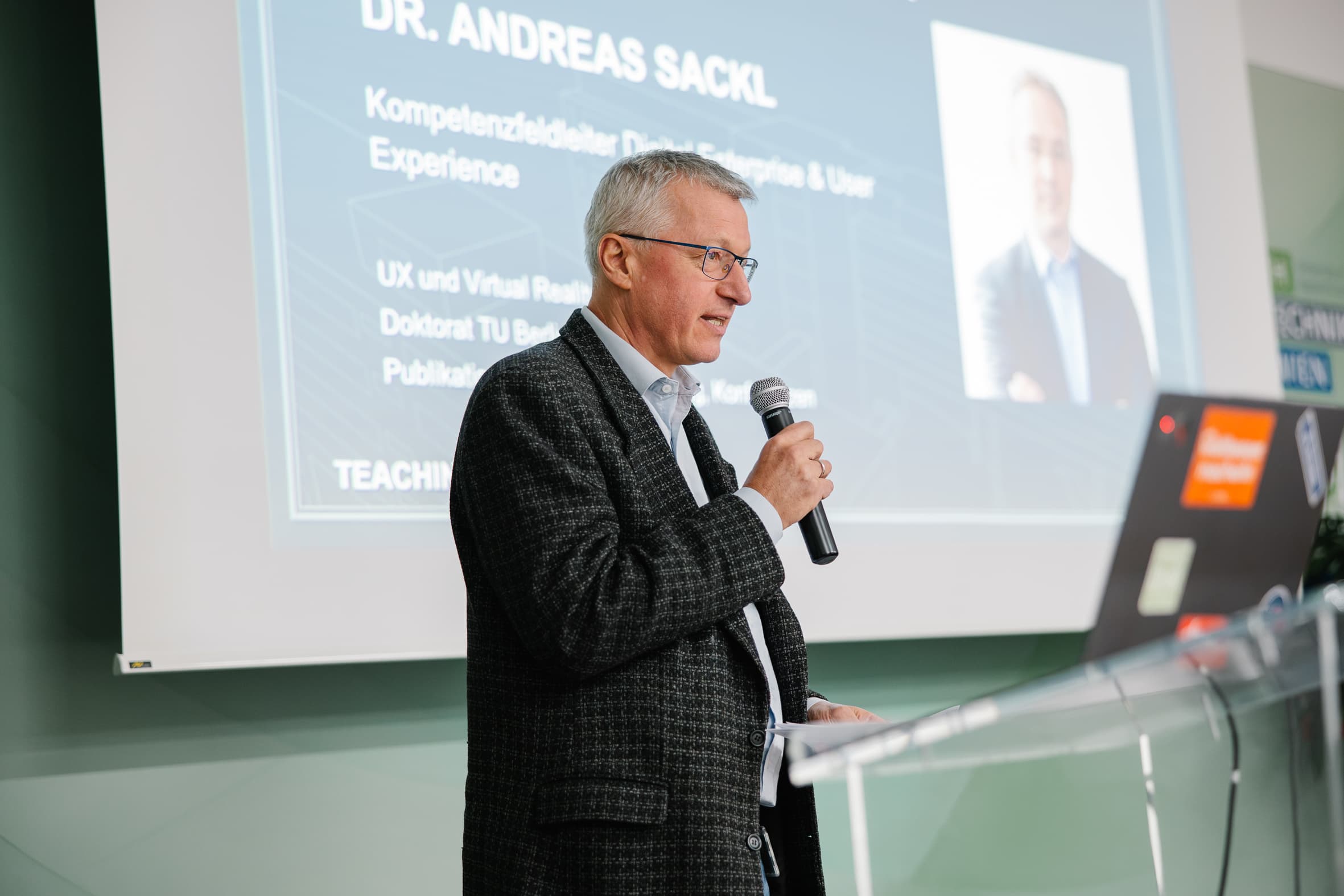 Dr. Andreas Sackl steht an einem Rednerpult und spricht in ein Mikrofon vor einer Leinwand, auf der sein Name und seine beruflichen Angaben zu sehen sind.