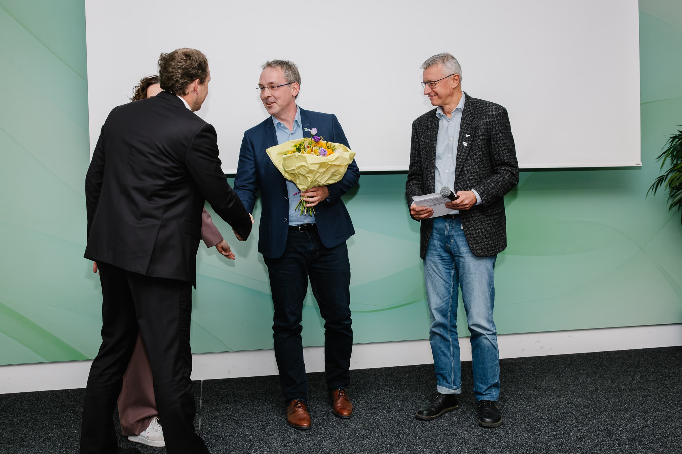 Ein Mann in einem blauen Blazer hält einen Blumenstrauß in der Hand, während er einem Mann in einem schwarzen Anzug die Hand schüttelt; daneben steht ein weiterer Mann mit einem Mikrofon in der Hand.