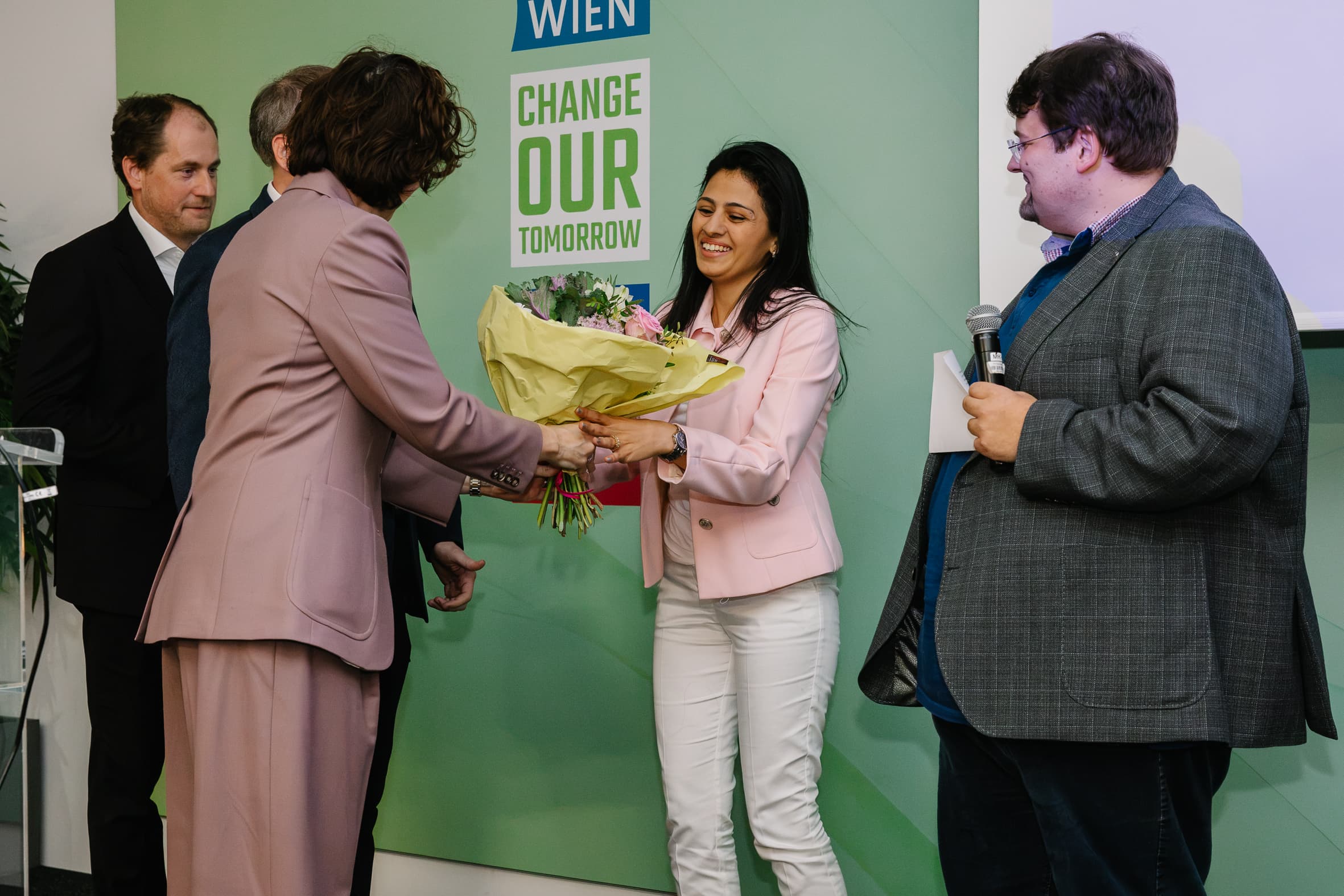 Eine Frau in einer rosa Jacke erhält von einer anderen Frau einen Blumenstrauß, während zwei Männer vor einem Schild mit der Aufschrift Change Our Tomorrow zusehen.