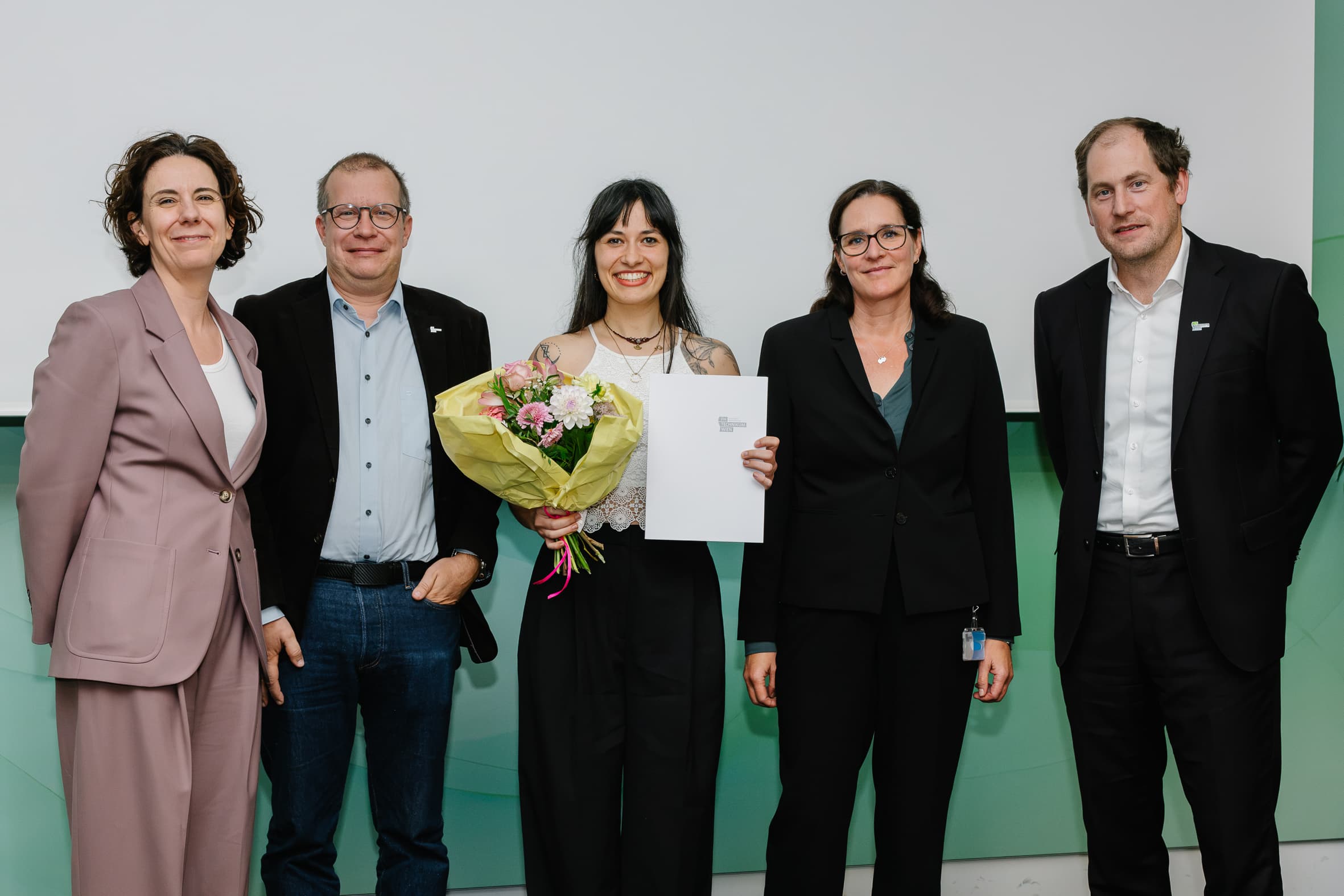 Fünf Personen stehen für ein Foto in einer Reihe, wobei die Frau in der Mitte einen Blumenstrauß und eine Urkunde in der Hand hält.