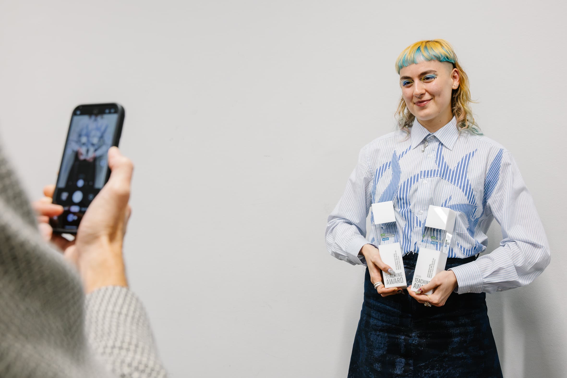 Eine Person mit blauen und gelben Haaren, die lächelnd fotografiert wird und zwei weiße, rechteckige Lehrpreise in den Händen hält.