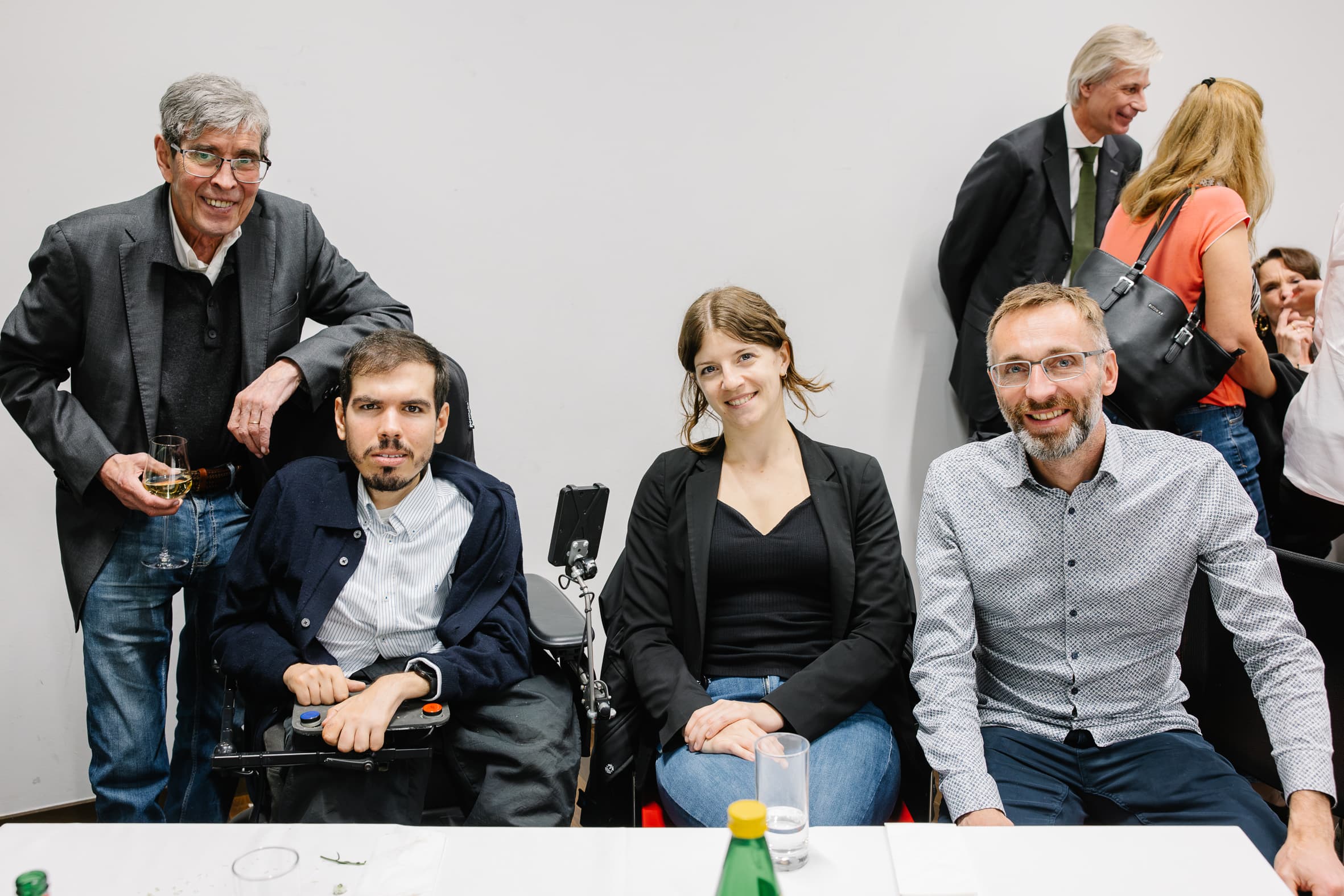 Vier Personen, die an einem Tisch für ein Foto posieren, darunter ein Mann im Rollstuhl; im Hintergrund sind weitere Gäste zu sehen.