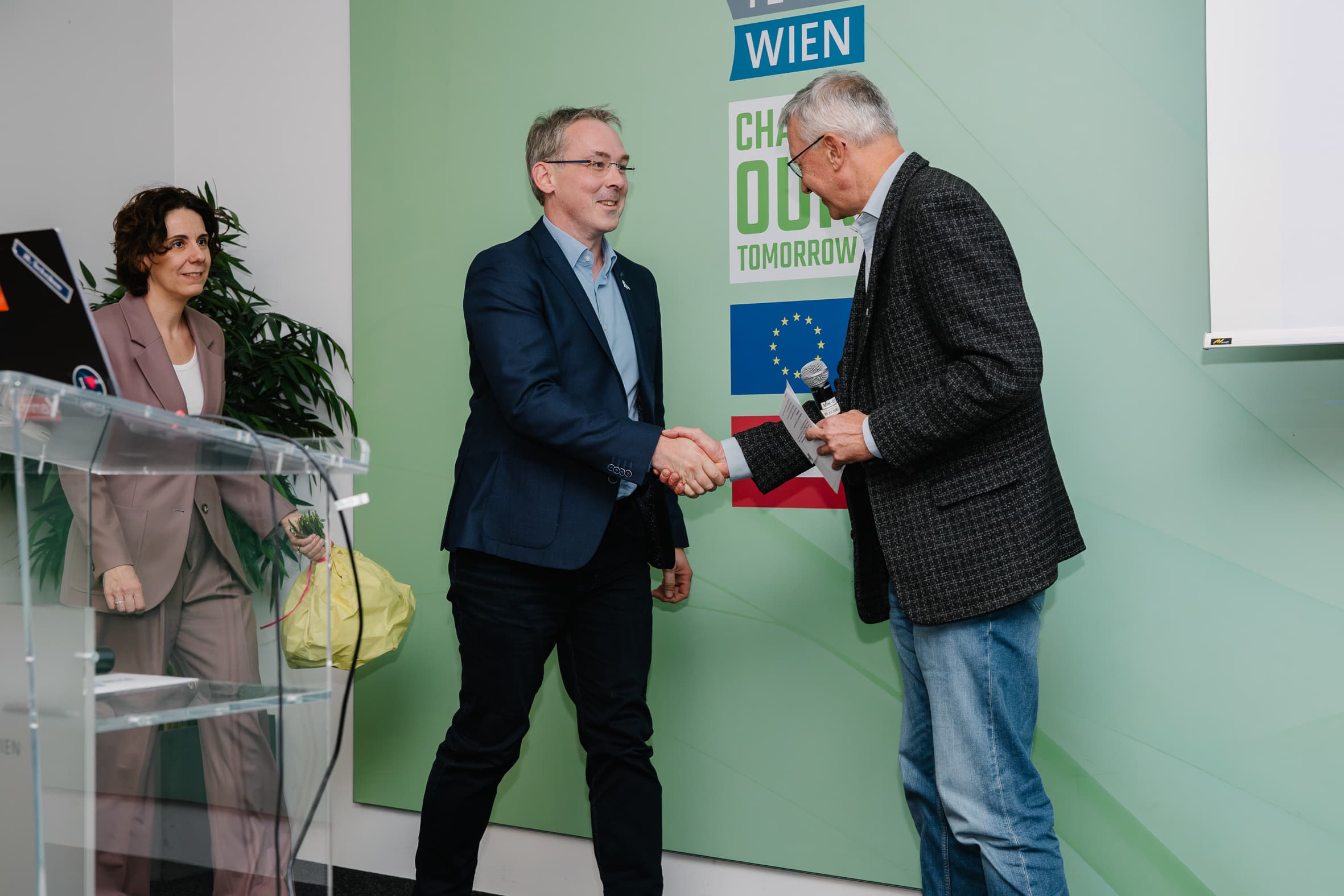 Zwei Männer geben sich auf der Bühne vor einer grünen Wand mit dem Logo der Europäischen Union und einem Text über Wien die Hand.