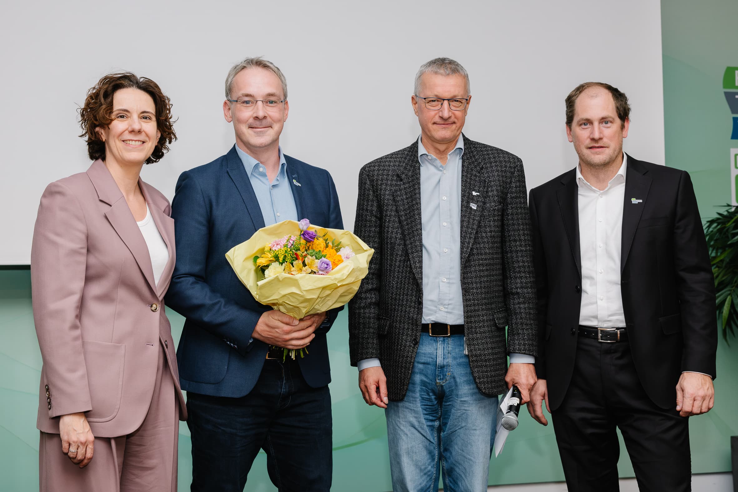 Vier Personen stehen in einer Reihe, wobei die zweite Person von links einen Blumenstrauß in der Hand hält.
