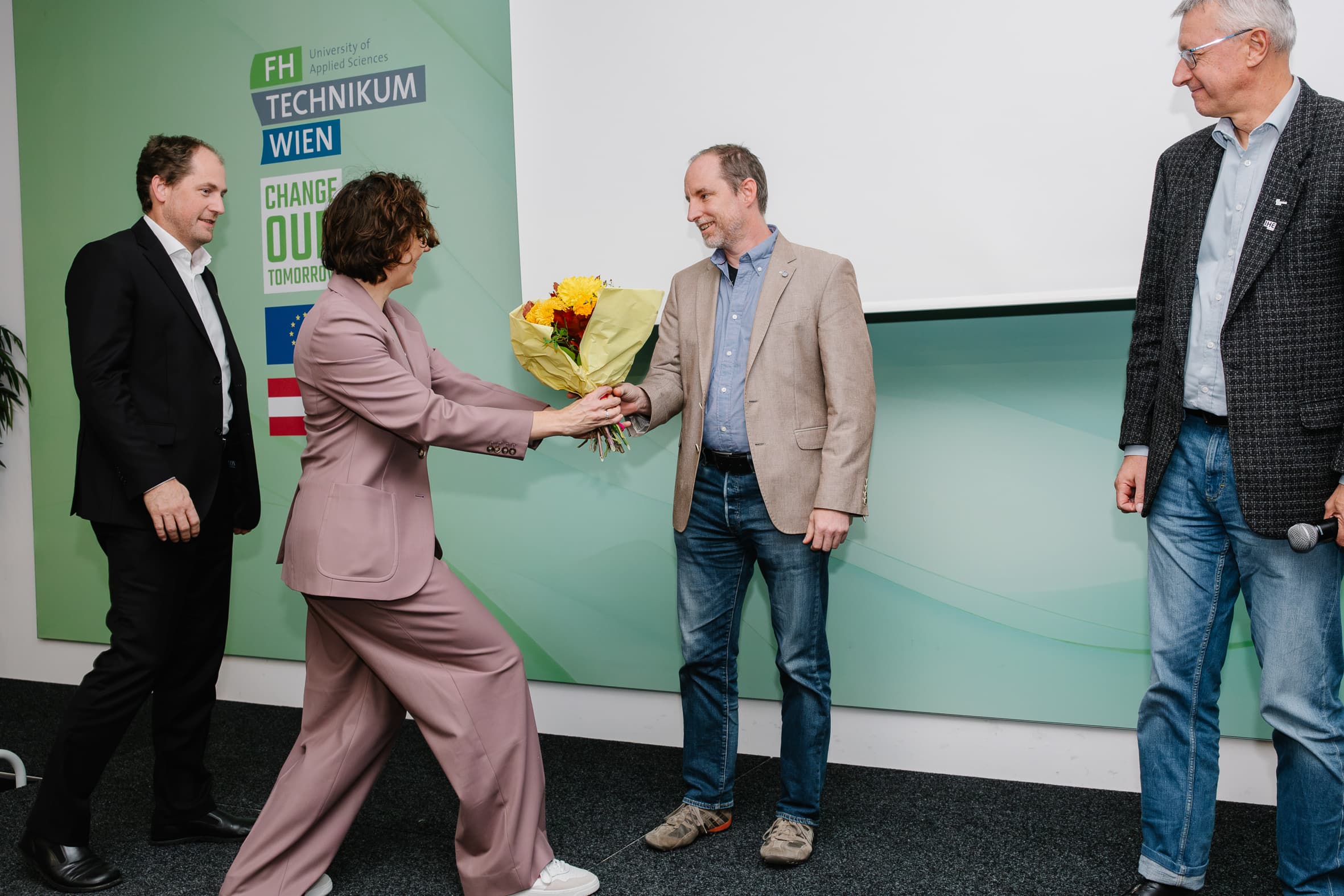 Eine Frau im Kostüm überreicht einem Mann in einem beigen Blazer einen Blumenstrauß, während zwei weitere Männer bei einer Veranstaltung vor einer Wand mit dem Logo des FH Technikum Wien zuschauen.