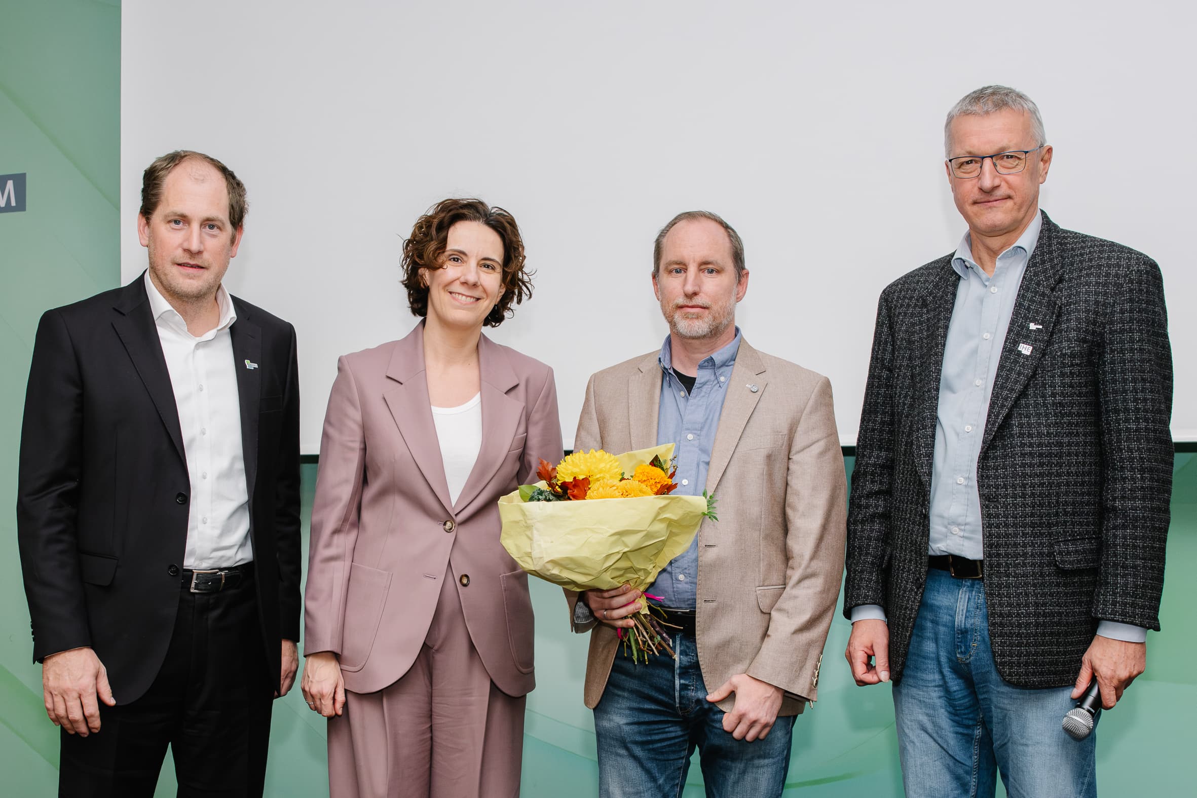 Vier Personen stehen in einer Reihe, wobei die zweite Person von links einen Blumenstrauß in der Hand hält und die Person ganz rechts ein Mikrofon.