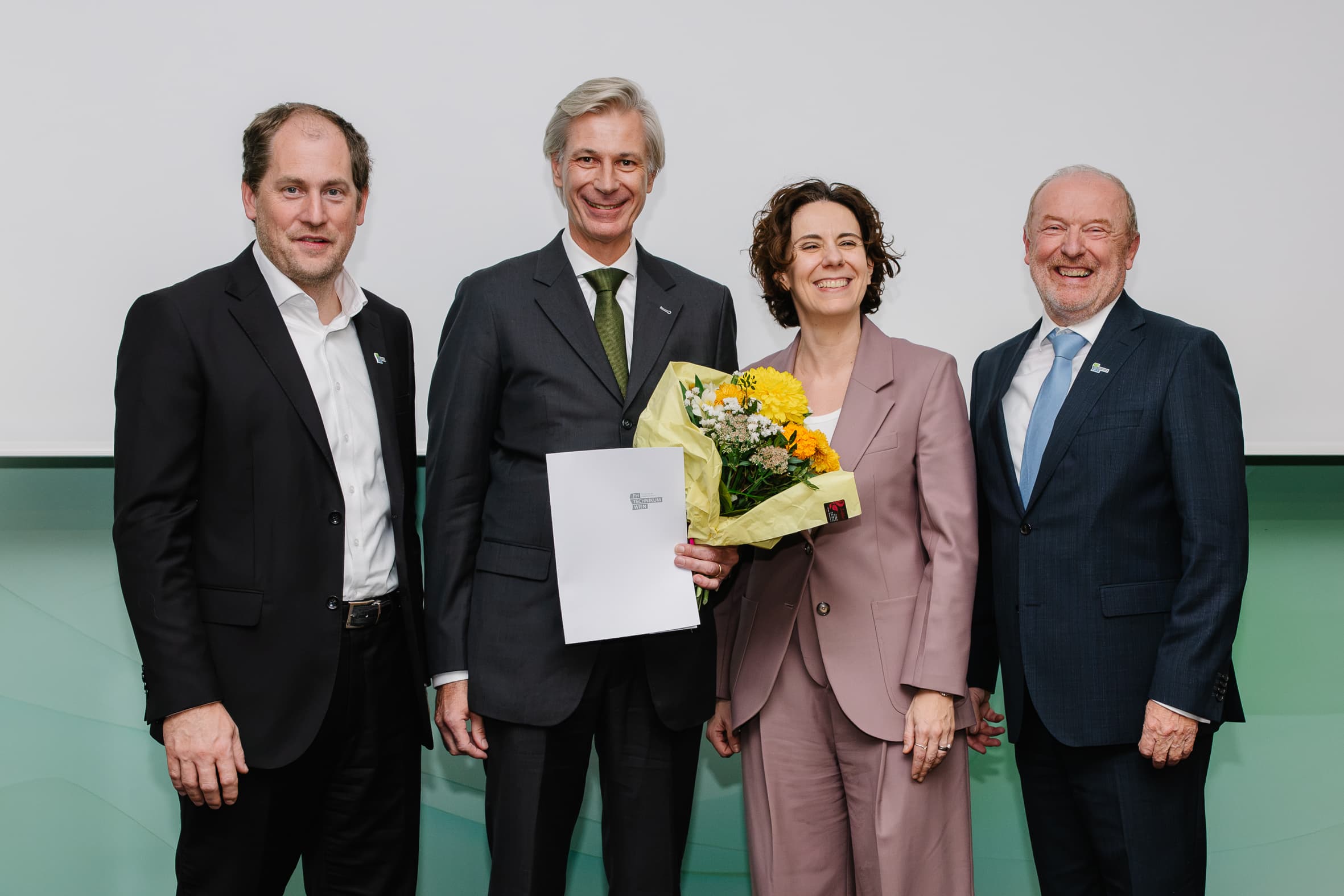 Vier Personen in Geschäftskleidung stehen beieinander, wobei die zweite Person von links einen Blumenstrauß und eine Urkunde in der Hand hält.
