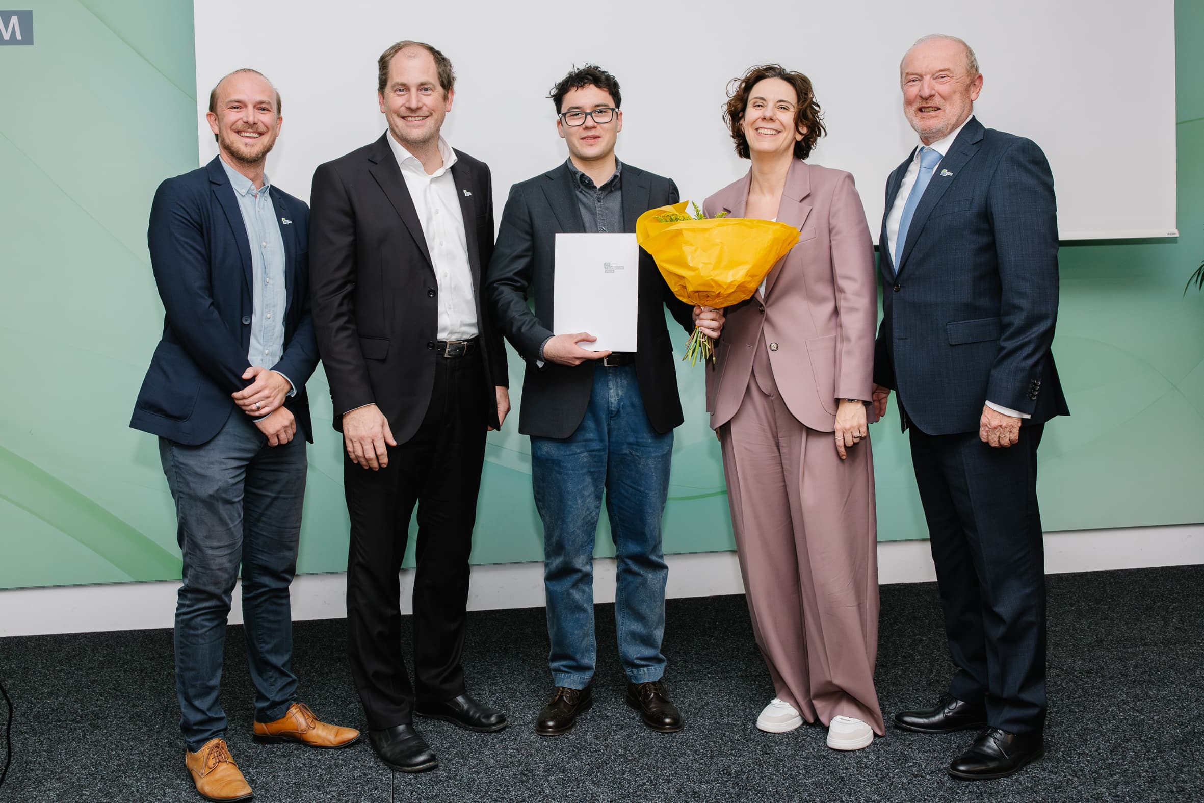 Fünf Personen stehen in einer Reihe auf einer Bühne; die Person in der Mitte hält eine Mappe in der Hand, und die Person rechts von ihr hält einen Strauß gelber Blumen.