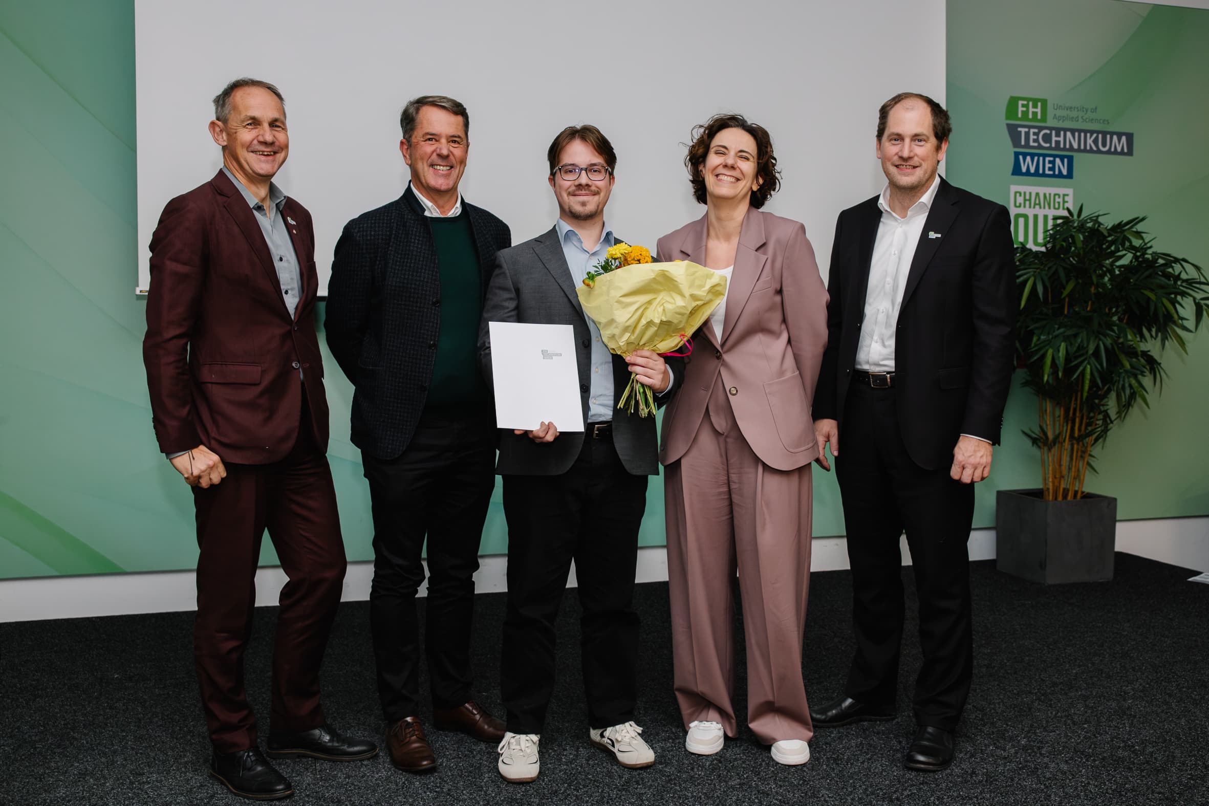 Fünf Personen stehen gemeinsam auf der Bühne, wobei die Person in der Mitte eine Urkunde und einen Blumenstrauß in den Händen hält.
