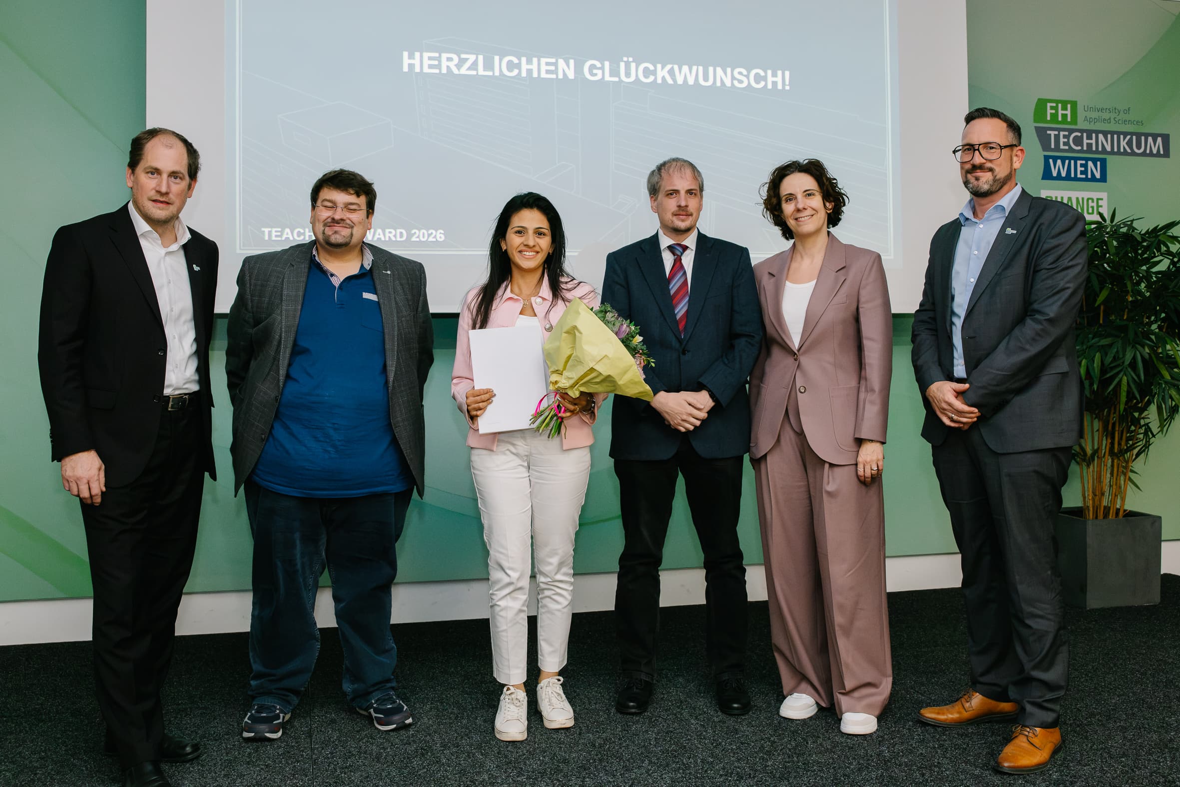 Fünf Personen stehen auf einer Bühne vor einer Leinwand, auf der HERZLICHEN GLÜCKWUNSCH! zu lesen ist; die Frau in der Mitte hält einen Blumenstrauß und ein Dokument in der Hand.
