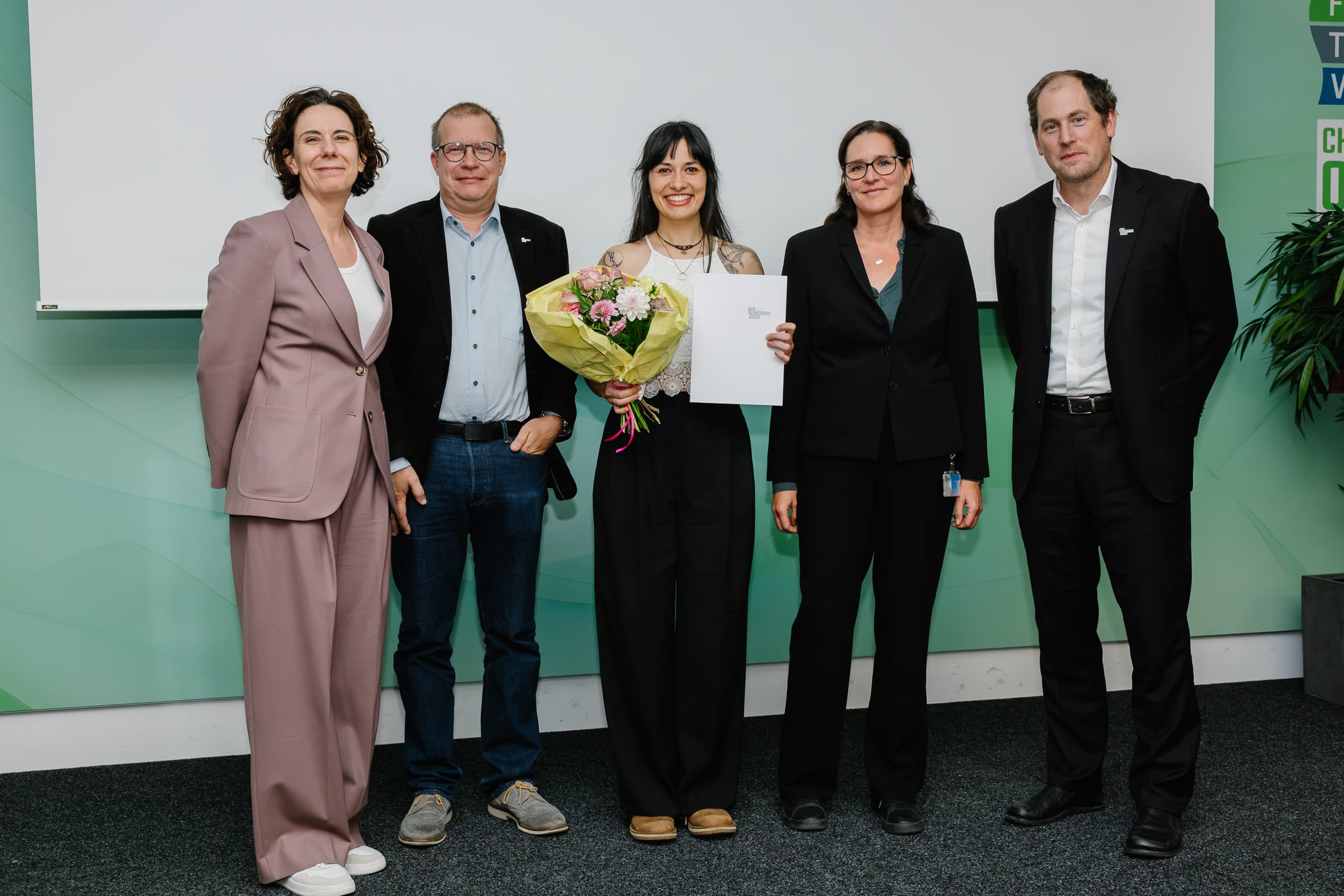 Fünf Personen stehen in einer Reihe auf einer Bühne, wobei die Person in der Mitte einen Blumenstrauß und eine Urkunde in der Hand hält.