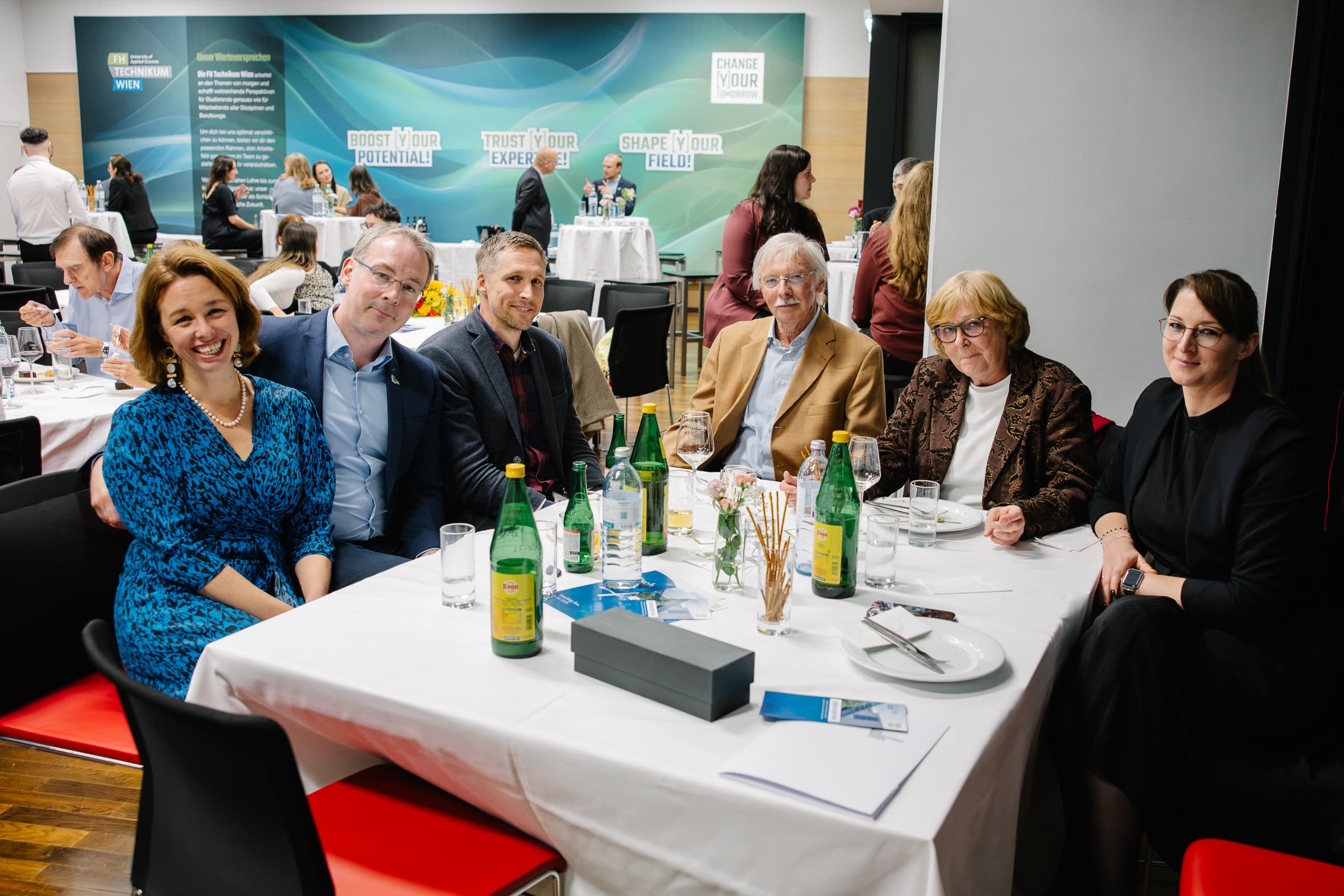 Sechs Personen sitzen bei einer Veranstaltung um einen Esstisch herum; im Hintergrund sind das Veranstaltungslogo und andere Teilnehmer zu sehen.