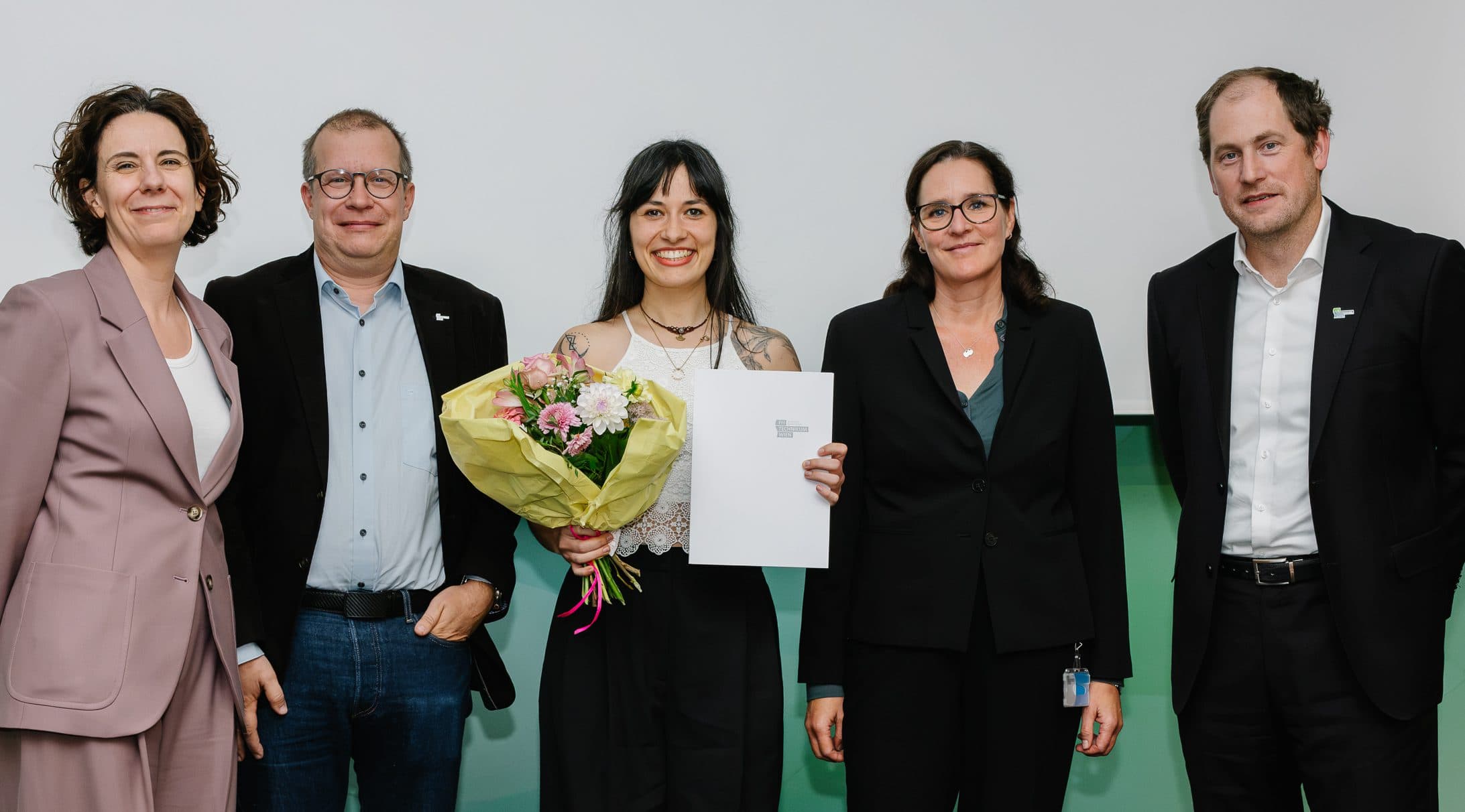 Fünf Personen stehen nebeneinander in einer Reihe, wobei die Frau in der Mitte einen Blumenstrauß und eine Urkunde in den Händen hält.