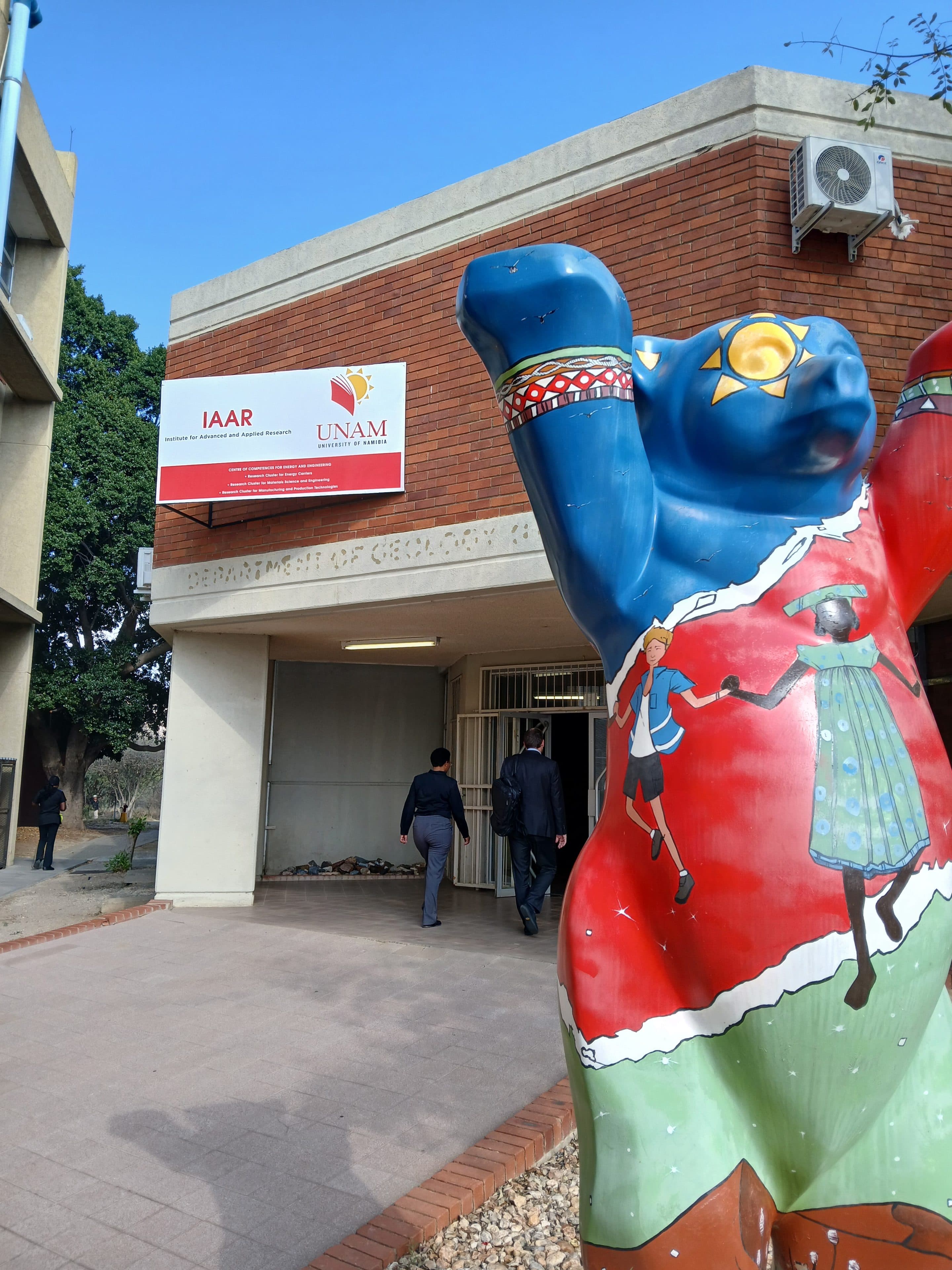 Vor dem Gebäude des Instituts für fortgeschrittene und angewandte Forschung der Universität Namibia steht eine große, farbenfrohe Bärenstatue, während zwei Personen auf den Eingang zugehen.