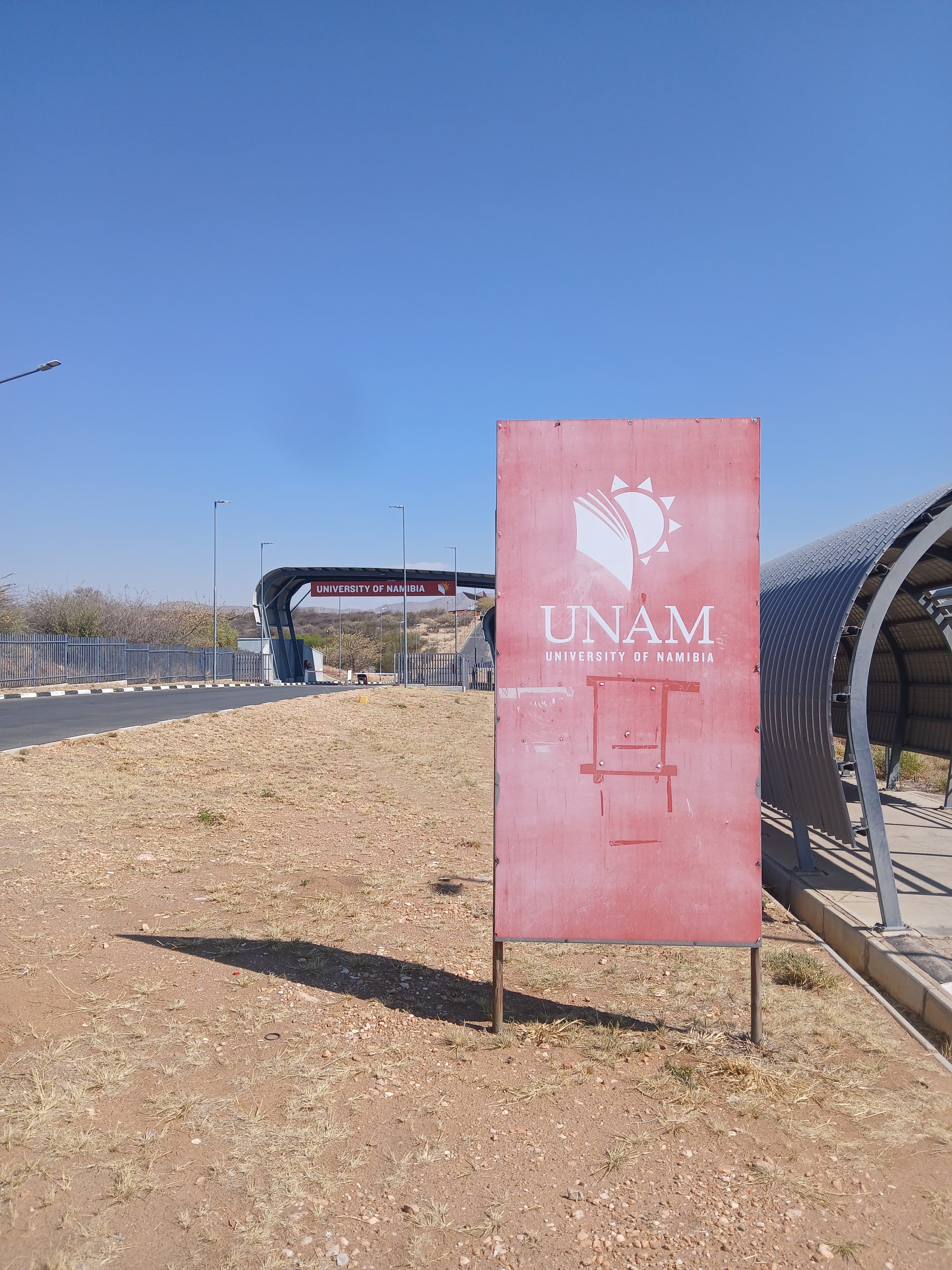 Ein Schild der Universität von Namibia steht auf einer trockenen, offenen Fläche in der Nähe eines Eingangstors zum Campus.