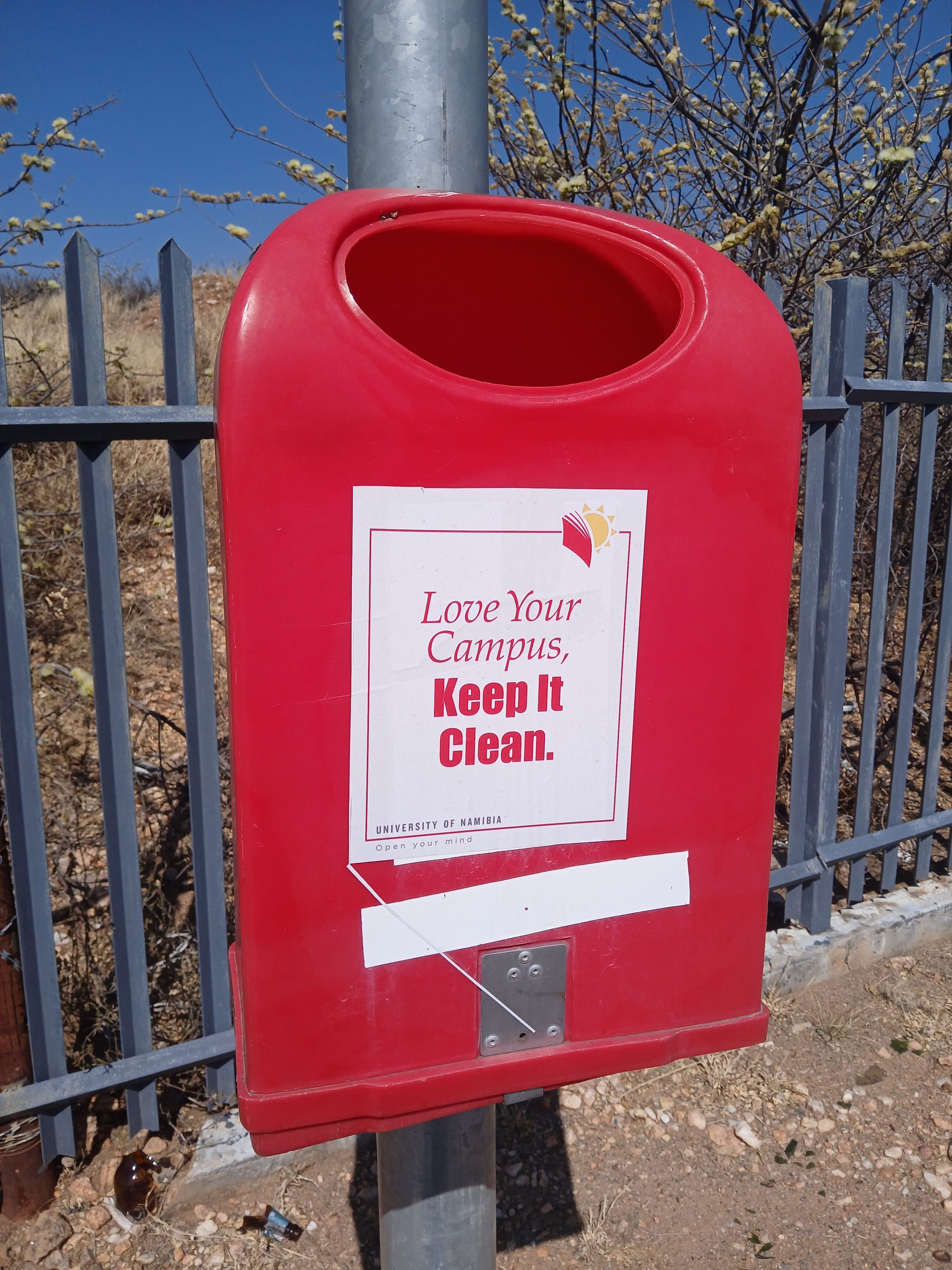 Rote Mülltonne mit einem Schild, auf dem steht: Liebt euren Campus, haltet ihn sauber. Universität von Namibia.